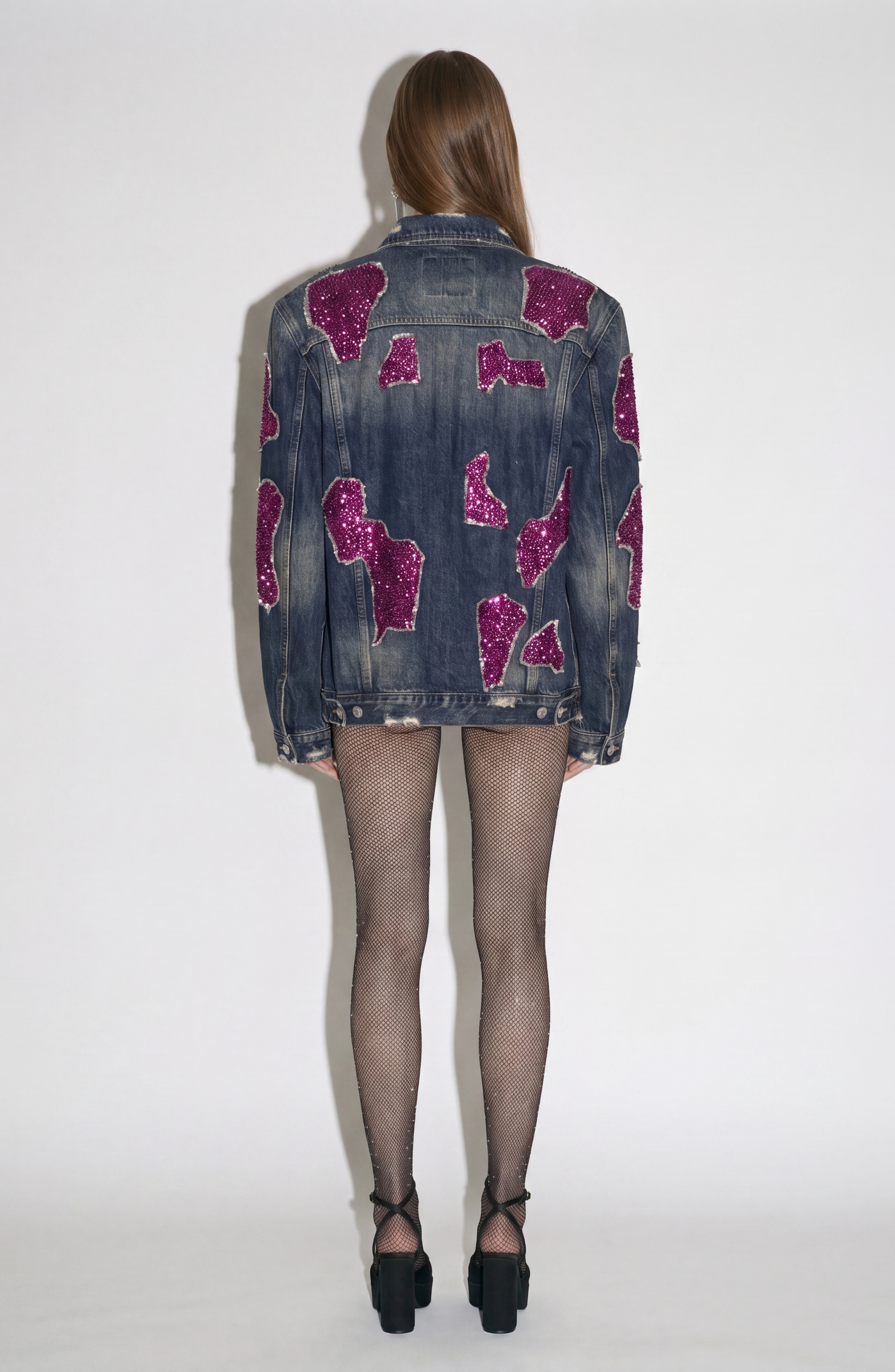 A person is shown from the back wearing the LOULOU Embellished Denim Jacket, featuring a vintage wash, irregular magenta sequin appliqués, and subtle padded shoulders. The look is styled with sheer fishnet tights and black high-heeled shoes against a plain white background.