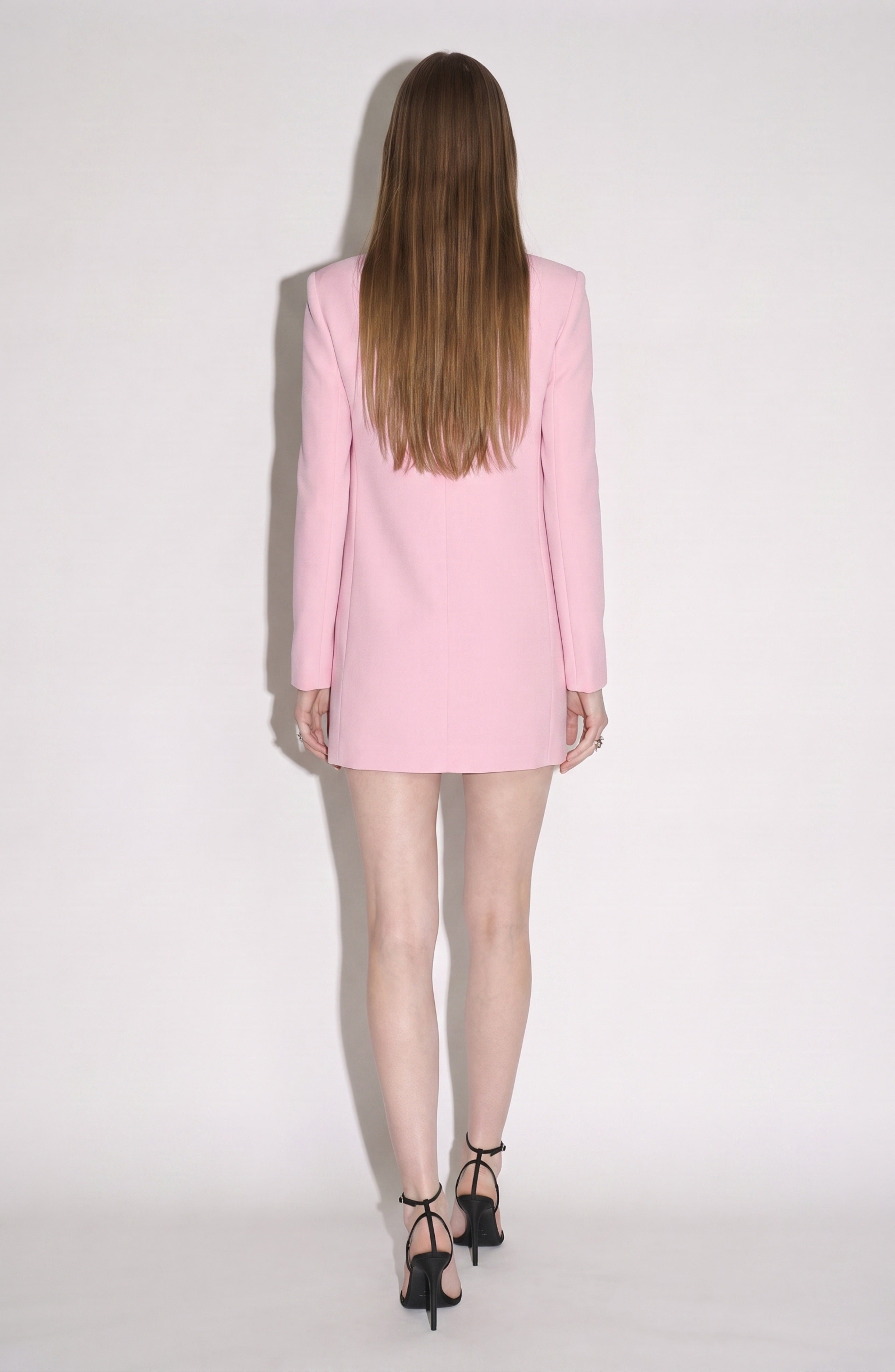 A woman with long brown hair stands facing away, wearing the LOULOU Embellished Mini Dress in powder pink, which features a tailored shift silhouette, paired with black high-heeled sandals against a plain white background.