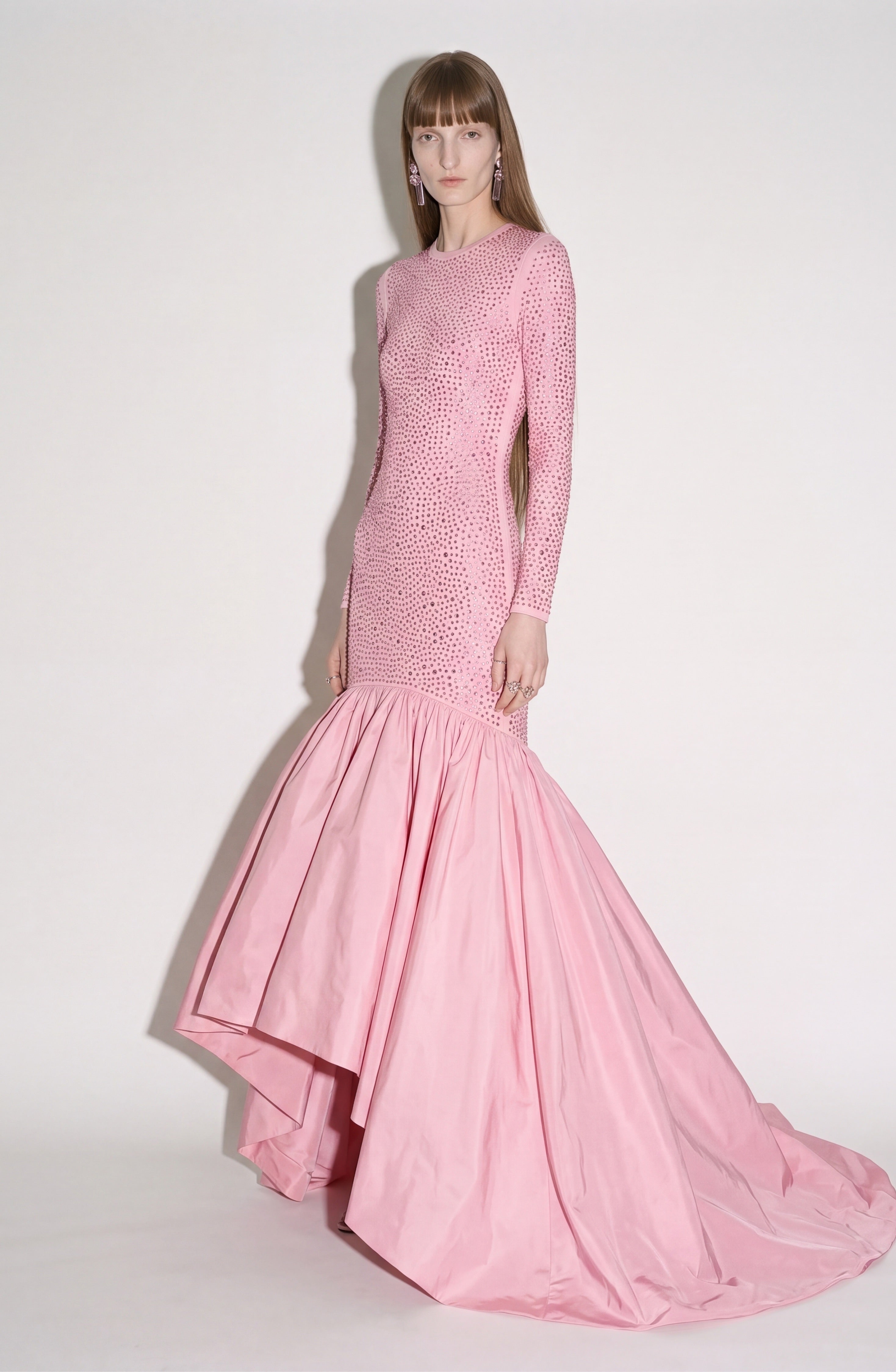 A woman poses against a plain light background wearing the LOULOU Embellished Taffeta Gown, featuring long sleeves, a fitted silhouette, crystal accents, and a dramatic flared mermaid skirt in light pink.