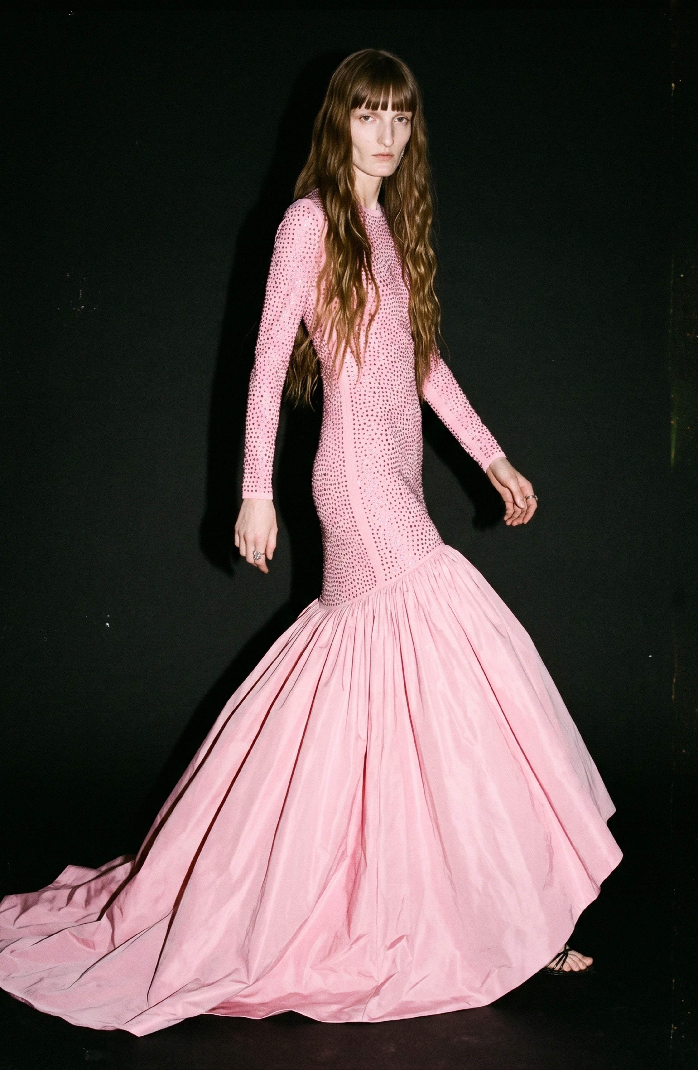A person with long, wavy brown hair wears the LOULOU Embellished Taffeta Gown—a fitted, long-sleeve pink evening dress featuring textured fabric and a voluminous high-low taffeta skirt—while standing against a black background.
