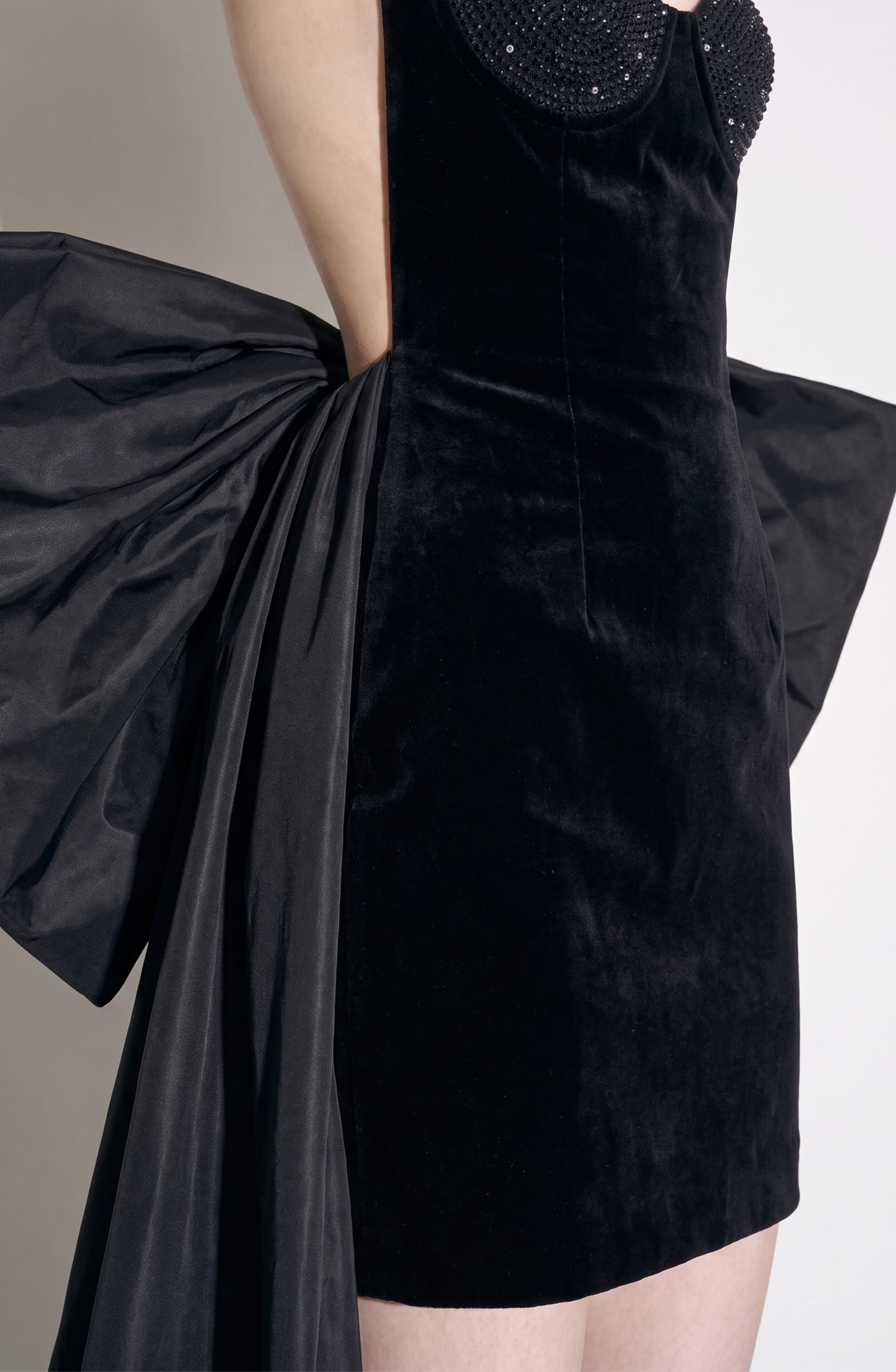 A person models the LOULOU Velvet Bow Mini Dress, featuring black velvet fabric and a statement side taffeta bow, standing against a plain backdrop with only the torso and upper legs in view.
