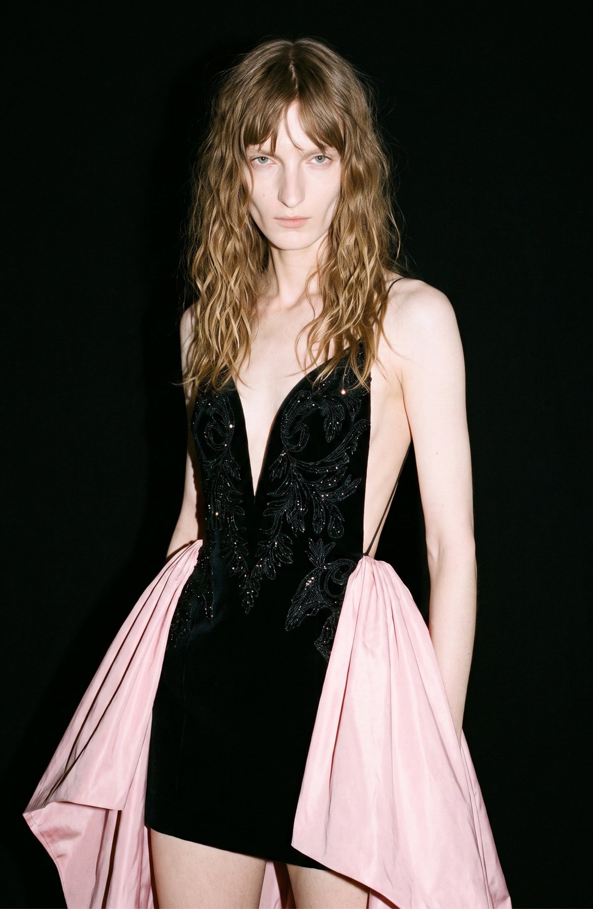 A person with wavy, light brown hair wears the LOULOU Embellished Velvet Mini Dress, a sleeveless black dress with a deep neckline, intricate designs, and a striking pink taffeta overskirt, standing against a black background.