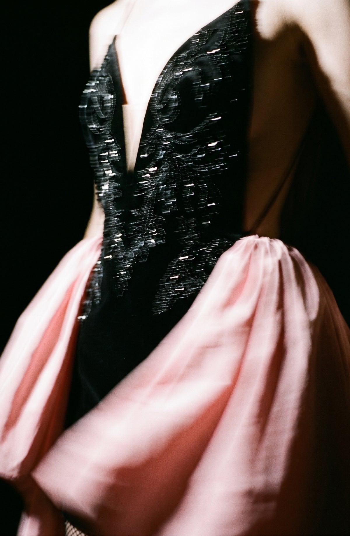 A person wearing the LOULOU Embellished Velvet Mini Dress—featuring black sequined velvet with intricate beadwork and a deep V-neck—paired with a flowing pink taffeta overskirt, photographed in dramatic lighting against a dark background.