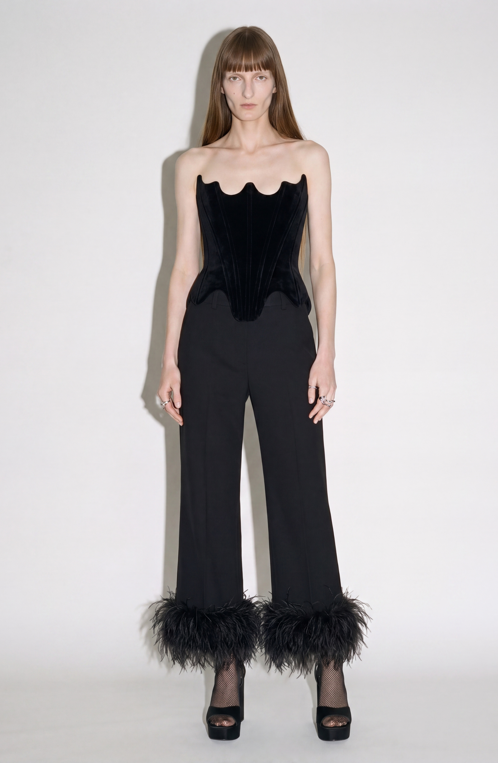 A person stands against a plain backdrop, wearing a black strapless velvet corset, LOULOU Feather Trim Tailored Trousers, open-toe heels, and minimal jewelry. The look is elegant and fashion-forward.