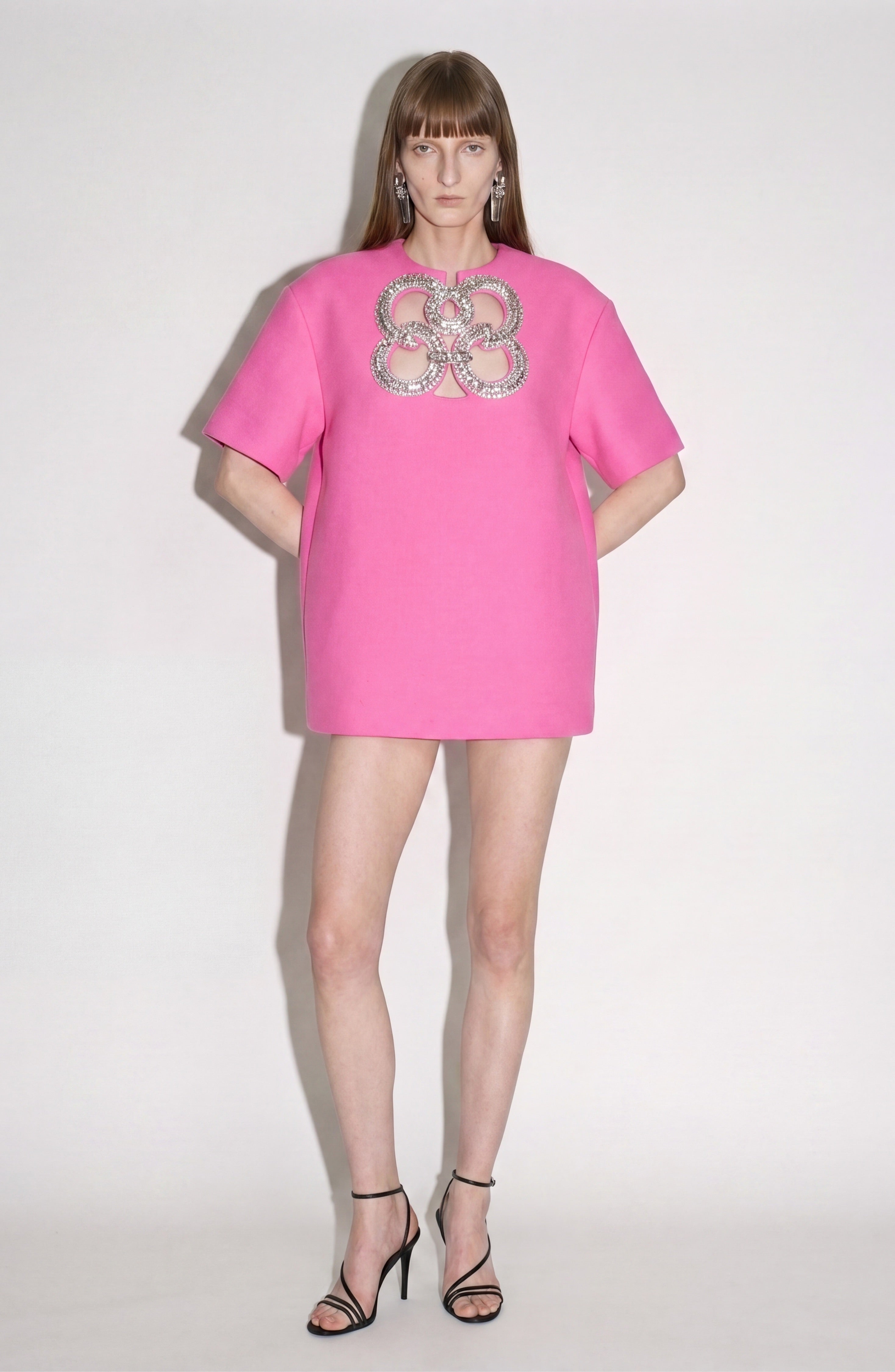 A woman stands against a plain background wearing the LOULOU Crystal Cutout Mini Dress in fuchsia, which features a circular jeweled cutout design on the chest, paired with black high-heeled sandals.