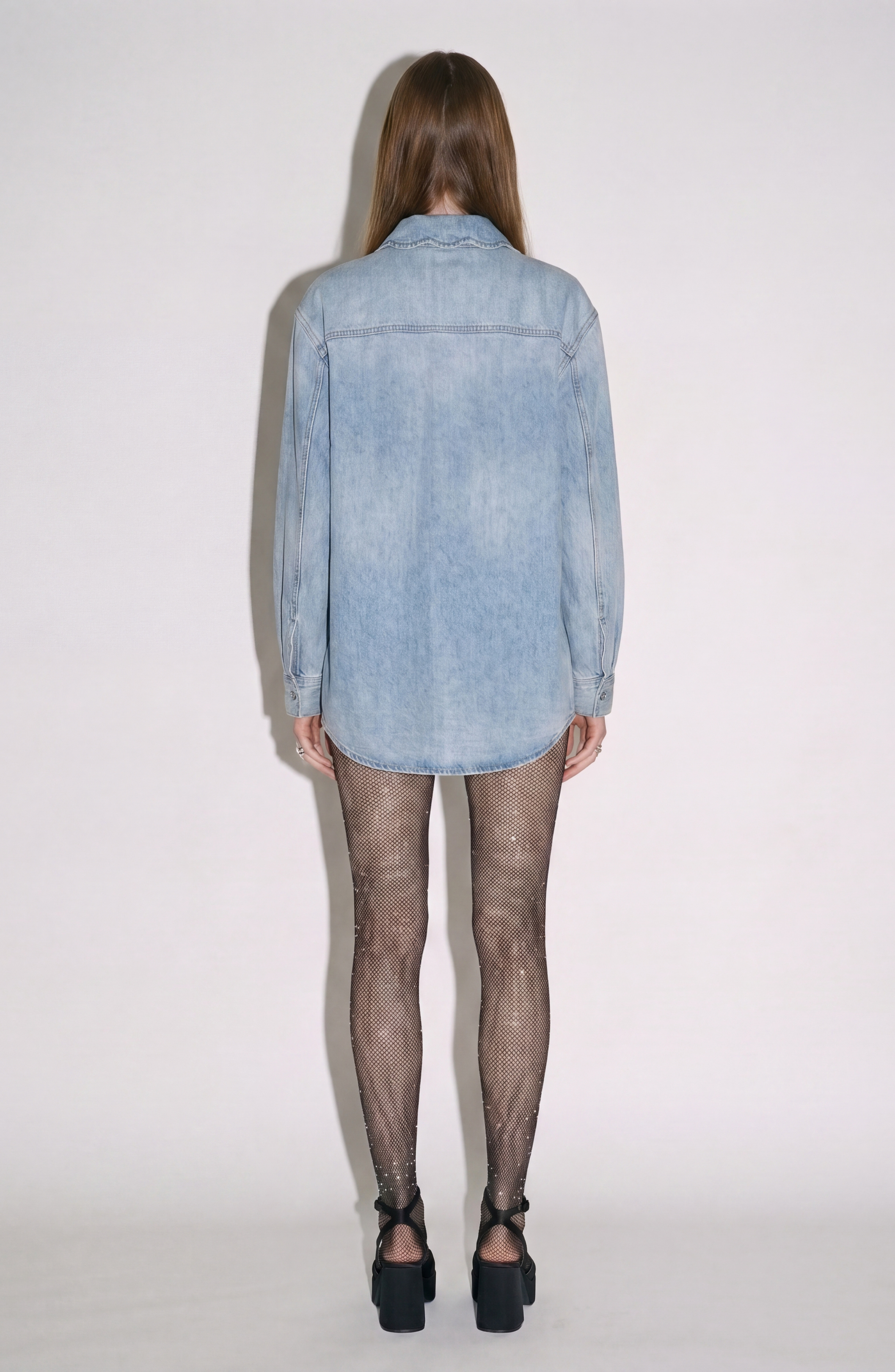 A person stands facing away against a plain white background, wearing the LOULOU Sequin Pocket Denim Shirt in a light wash, paired with black fishnet tights and black high-heeled shoes.