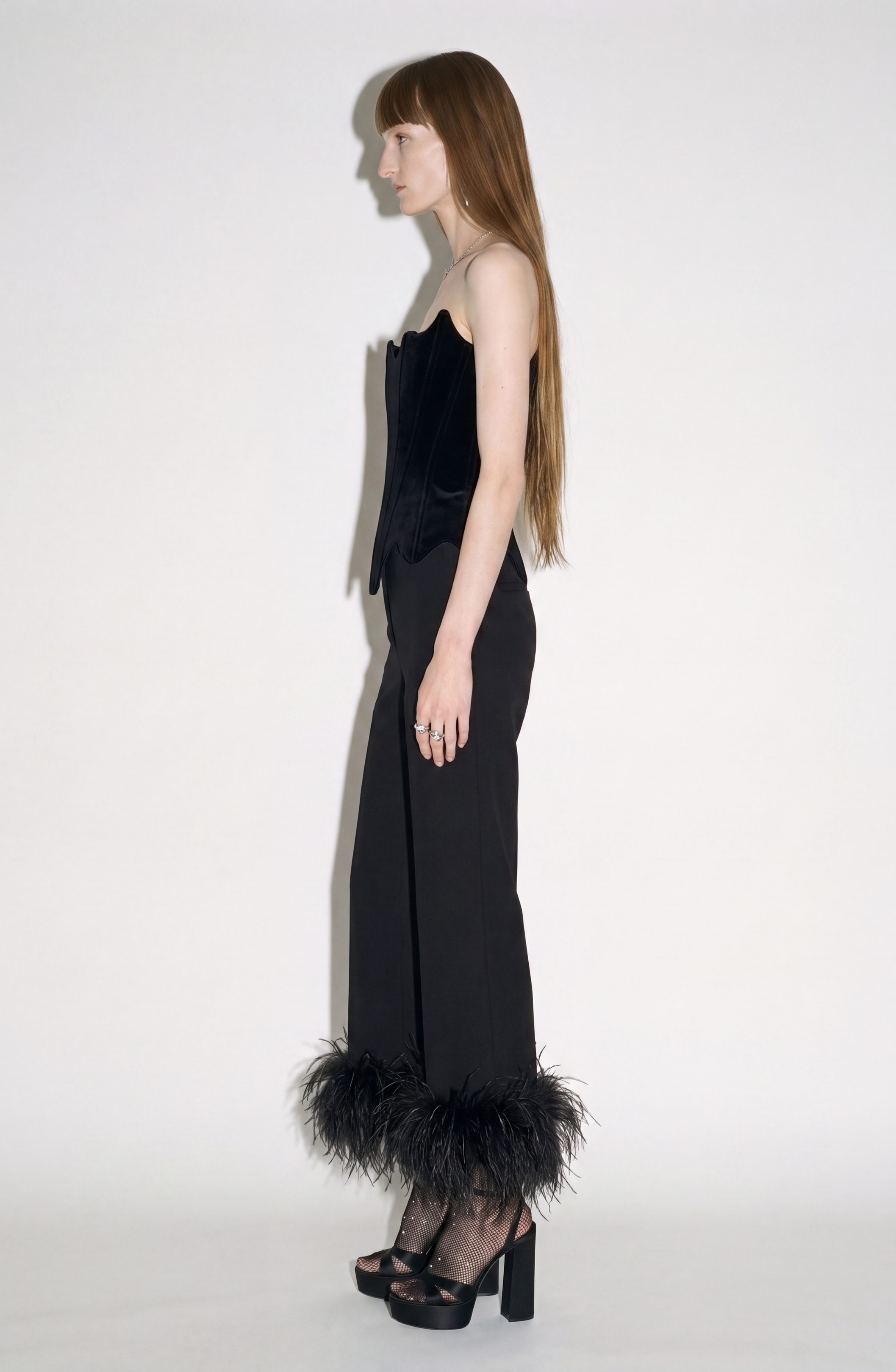 A model with long brown hair poses in profile against a white background. She wears the LOULOU Velvet Architectural Corset, a black velvet piece with an architectural neckline and lace-up back, paired with matching black trousers featuring feather trim at the hem, and black platform heels styled with fishnet socks.