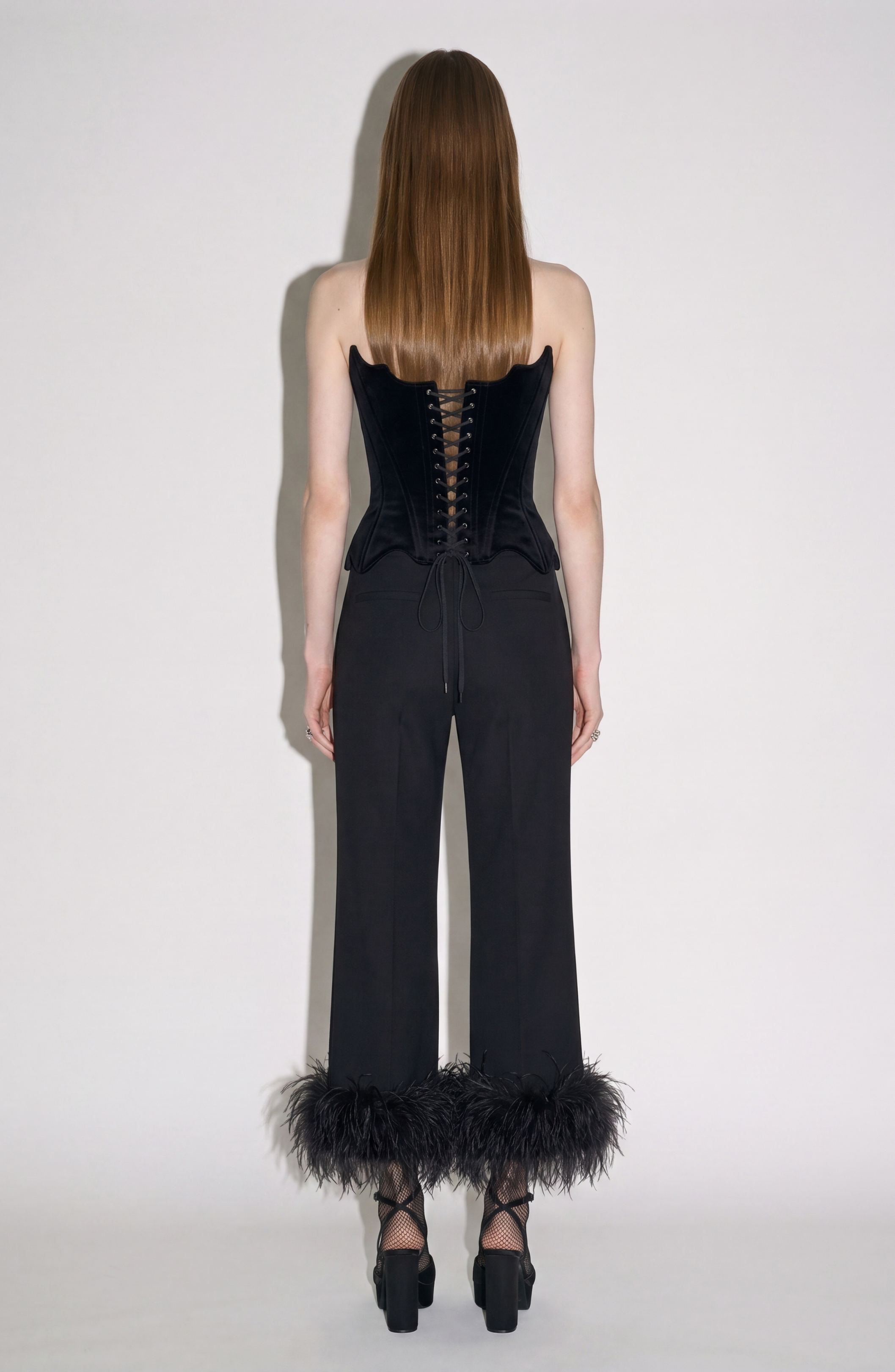 A woman stands facing away, wearing the LOULOU Velvet Architectural Corset featuring a dramatic lace-up back, paired with black pants with feather cuffs and platform shoes. She has straight brown hair, and the background is plain white.
