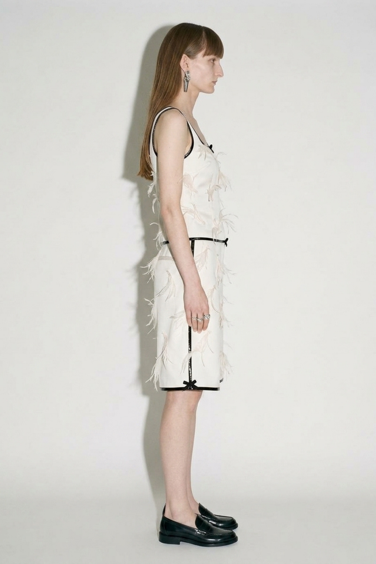 A woman with straight brown hair in profile wears a sleeveless light top and LOULOU Applique Bermuda Shorts—ivory with feather appliqué, black trim—and black loafers against a plain background.