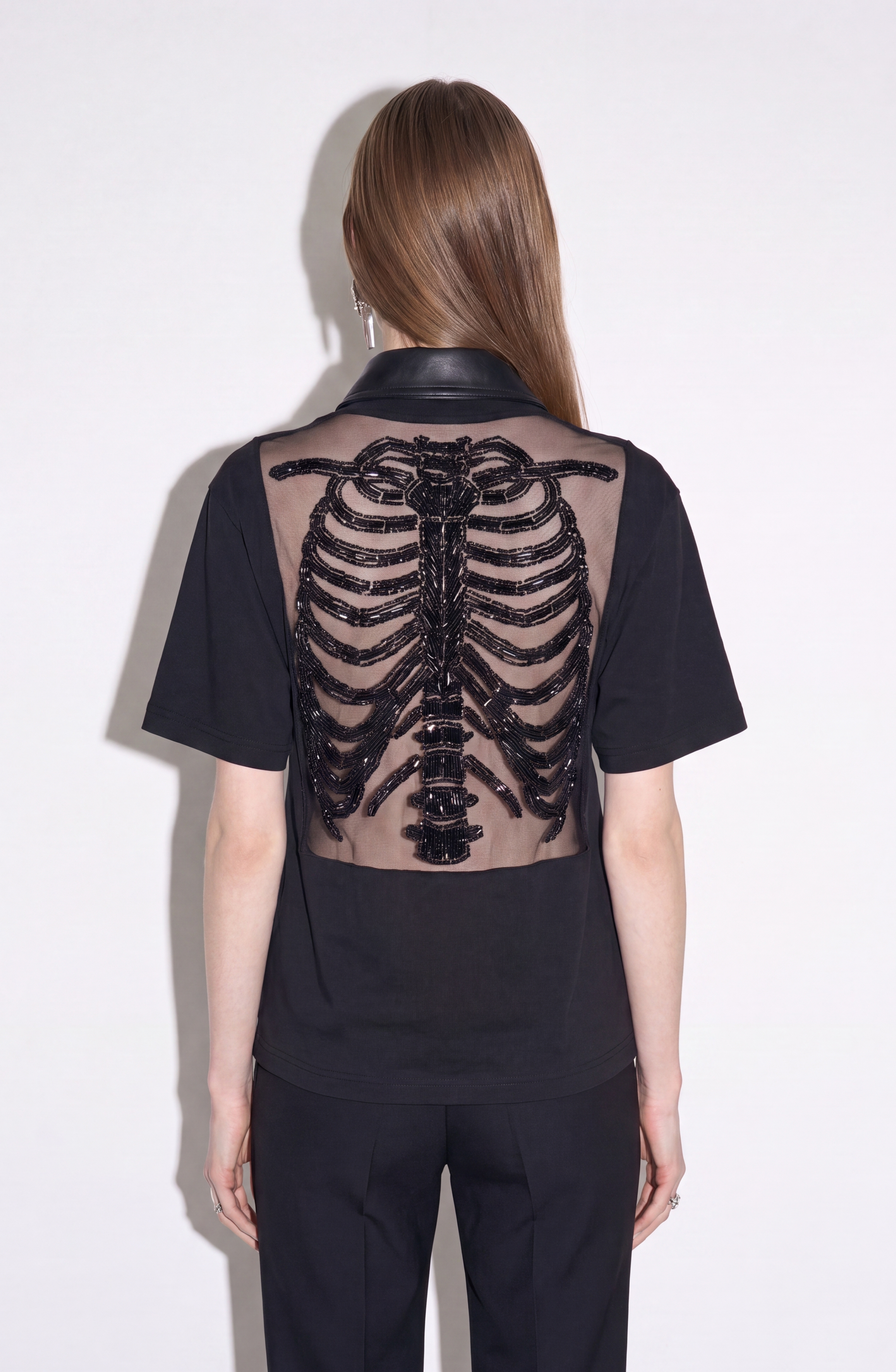 A person wearing the LOULOU Beaded Skeleton Polo Top in jet black, featuring a sheer back panel with a hand-beaded skeleton motif, stands against a plain light background.