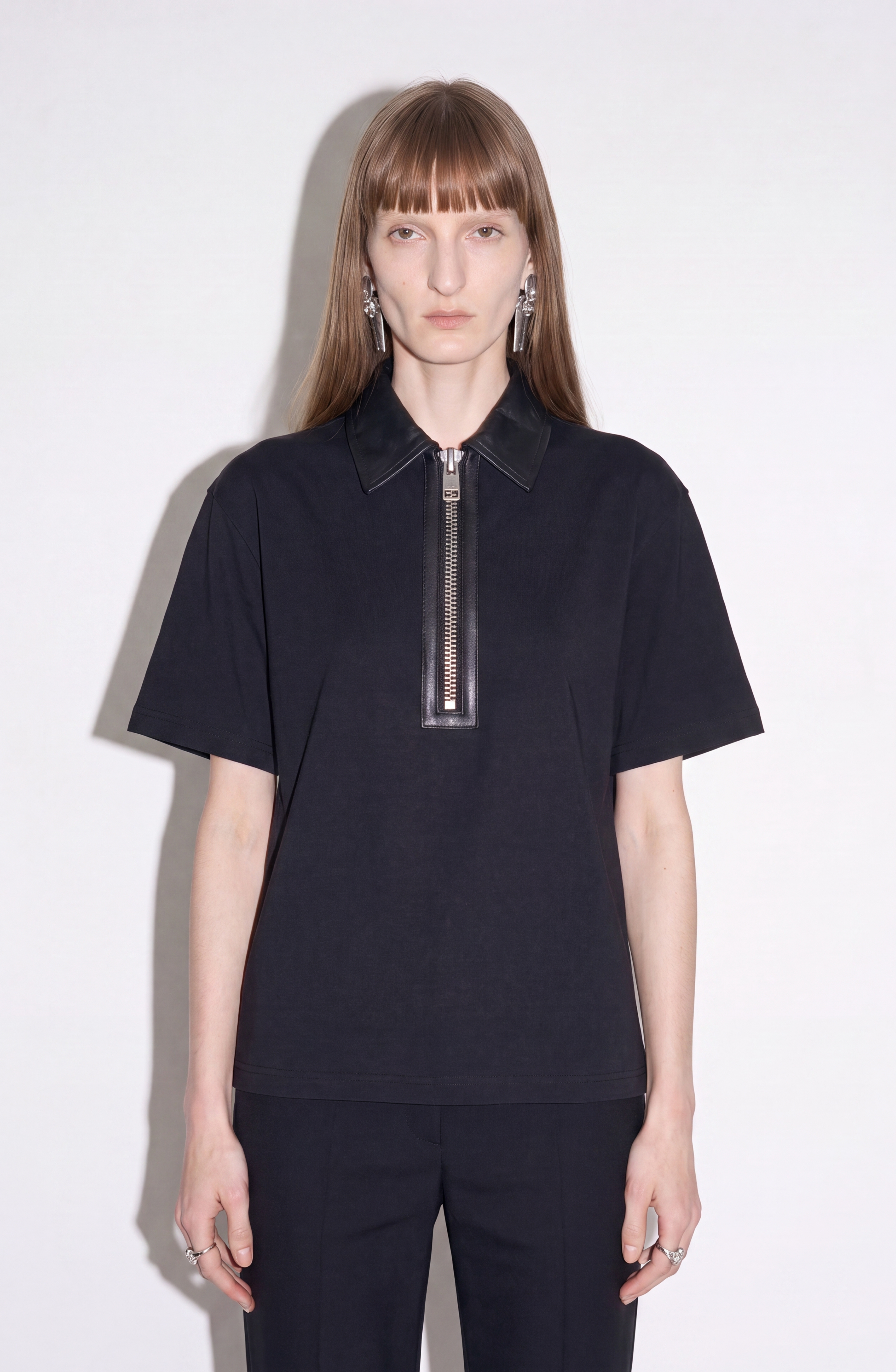 A person with long, straight hair wears the LOULOU Beaded Skeleton Polo Top—jet black with a large zippered leather collar—paired with black pants, standing against a plain white background.