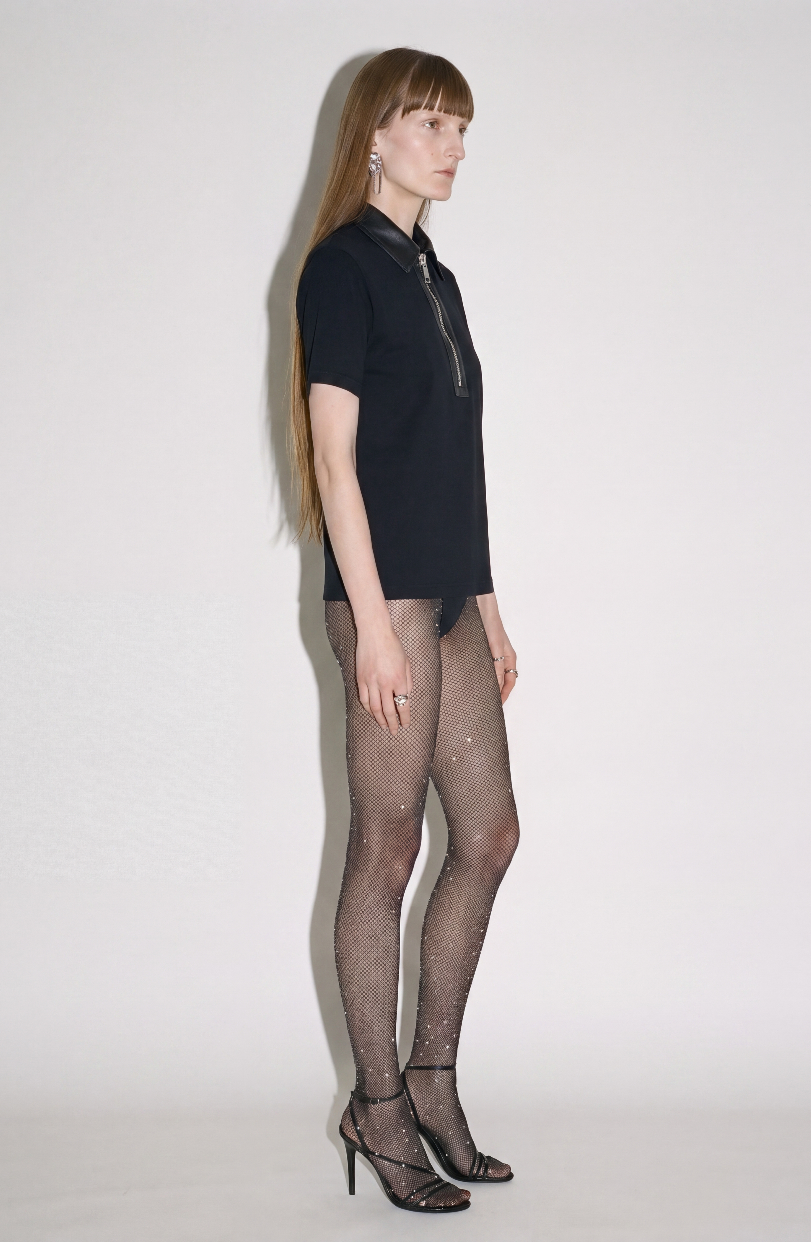 A person stands in profile against a plain white background, wearing the LOULOU Beaded Skeleton Polo Top—a jet black polo featuring a sleek leather collar and hand-beaded skeleton motif—styled with sheer embellished tights, black high-heeled sandals, statement earrings, and long straight hair.