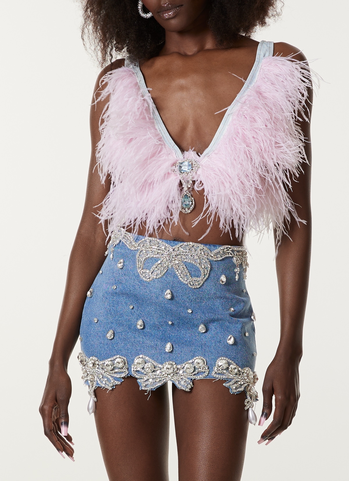 A woman wears a light purple feathered crop top with a deep V-neck, paired with the LOULOU Belle Antoinette Skirt—a blue piece adorned with pearls, rhinestones, and silver beading for a glamorous, embellished look.