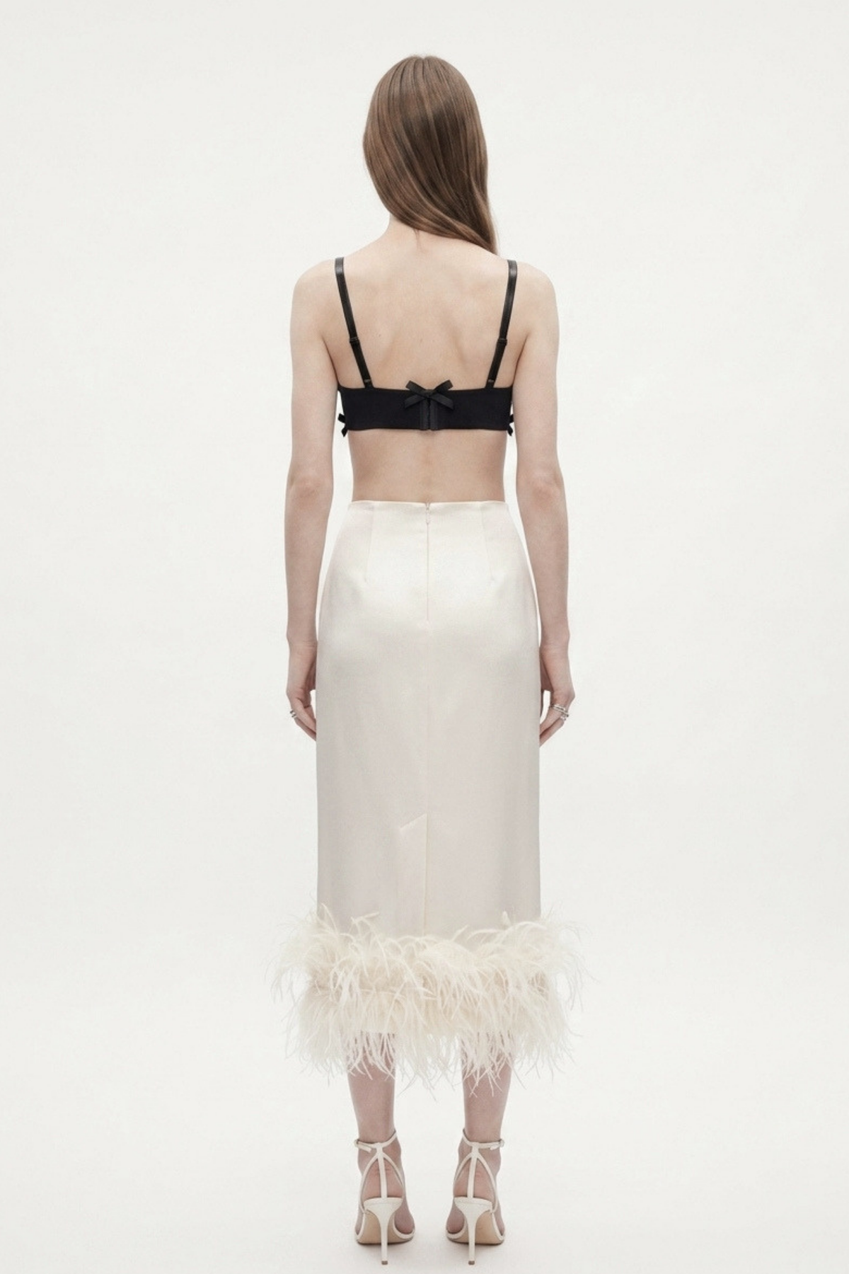 A woman stands with her back to the camera, wearing the LOULOU Black Bow Appliqué Bralette Top, a high-waisted white skirt with a feather-trimmed hem, and white strappy heels. Her straight hair is loose against a plain white background.
