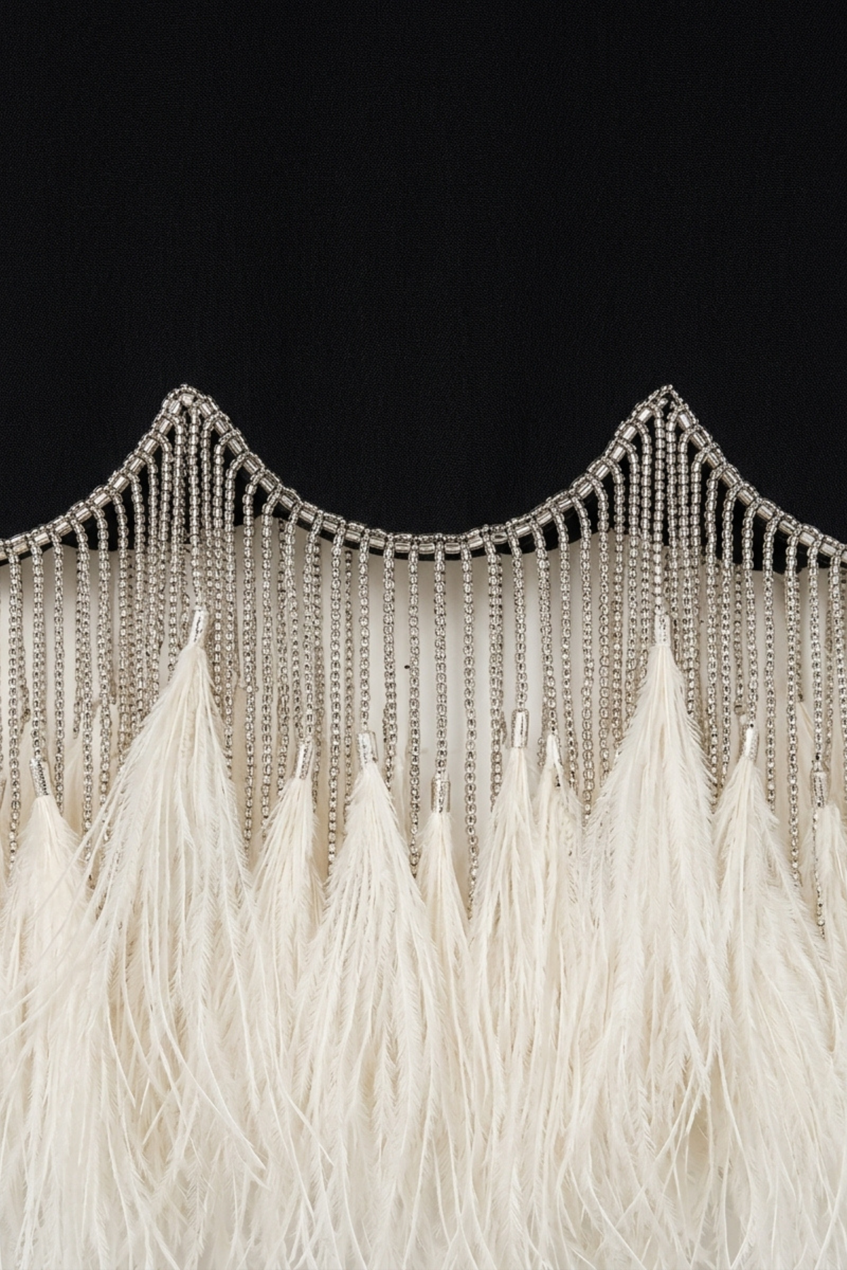 Close-up of the LOULOU Black Crystal Feather Long Sleeve Mini Dress, showing silver chains in wave patterns above soft white feather fringes, all set against a black background.