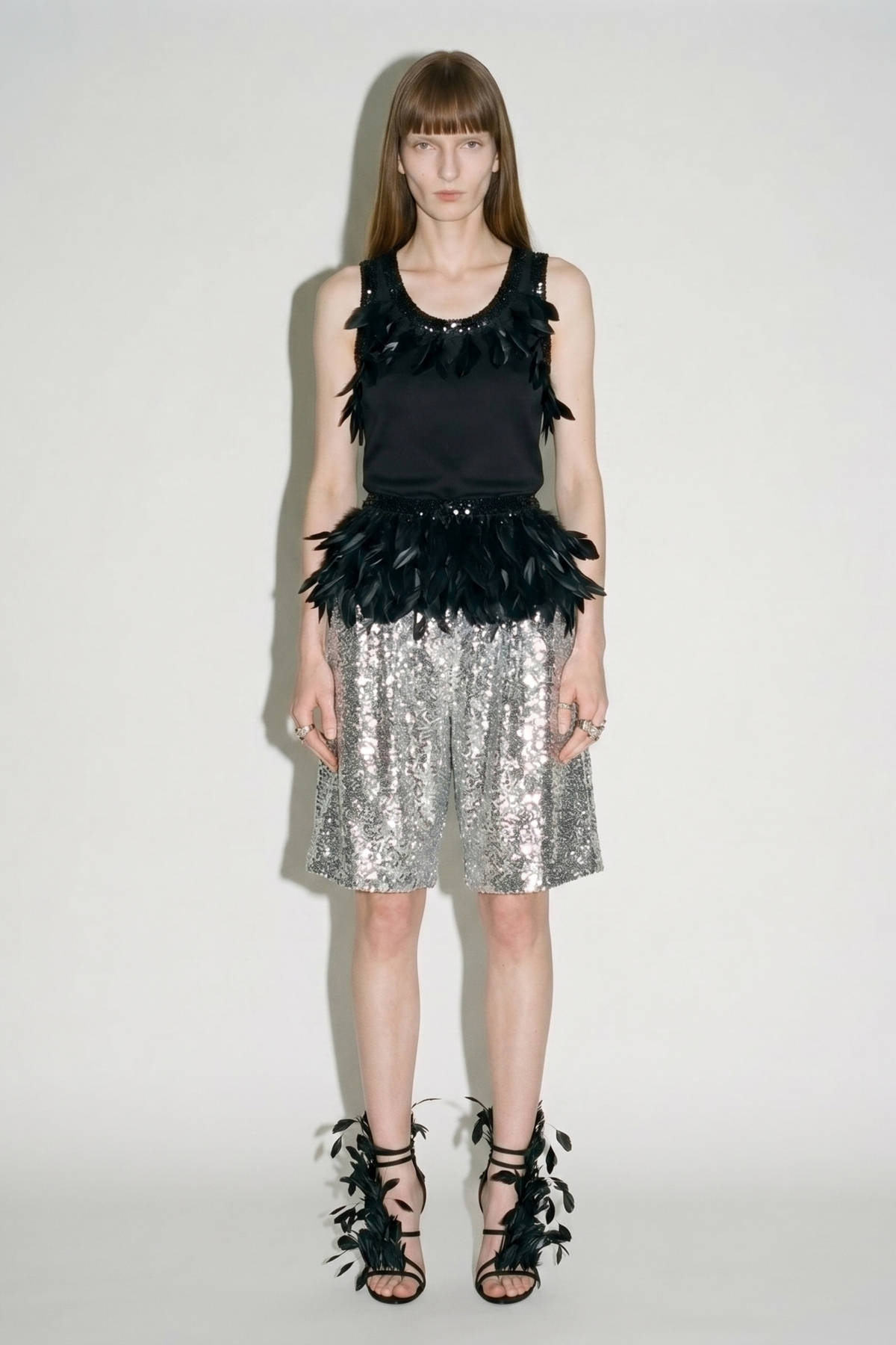 Model wearing the LOULOU Black Feather Sequin Peplum Top against a plain background, styled with silver sequined shorts and black feathered sandals.