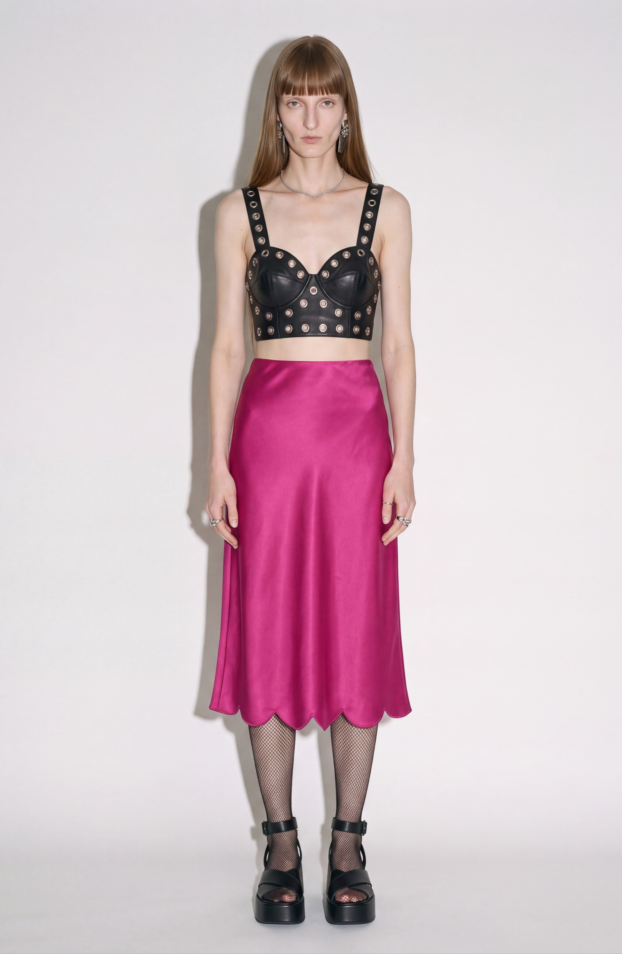 A woman stands against a white background wearing the LOULOU Black Leather Top with metal eyelets, paired with a bright pink satin midi skirt, fishnet stockings, and black platform sandals.