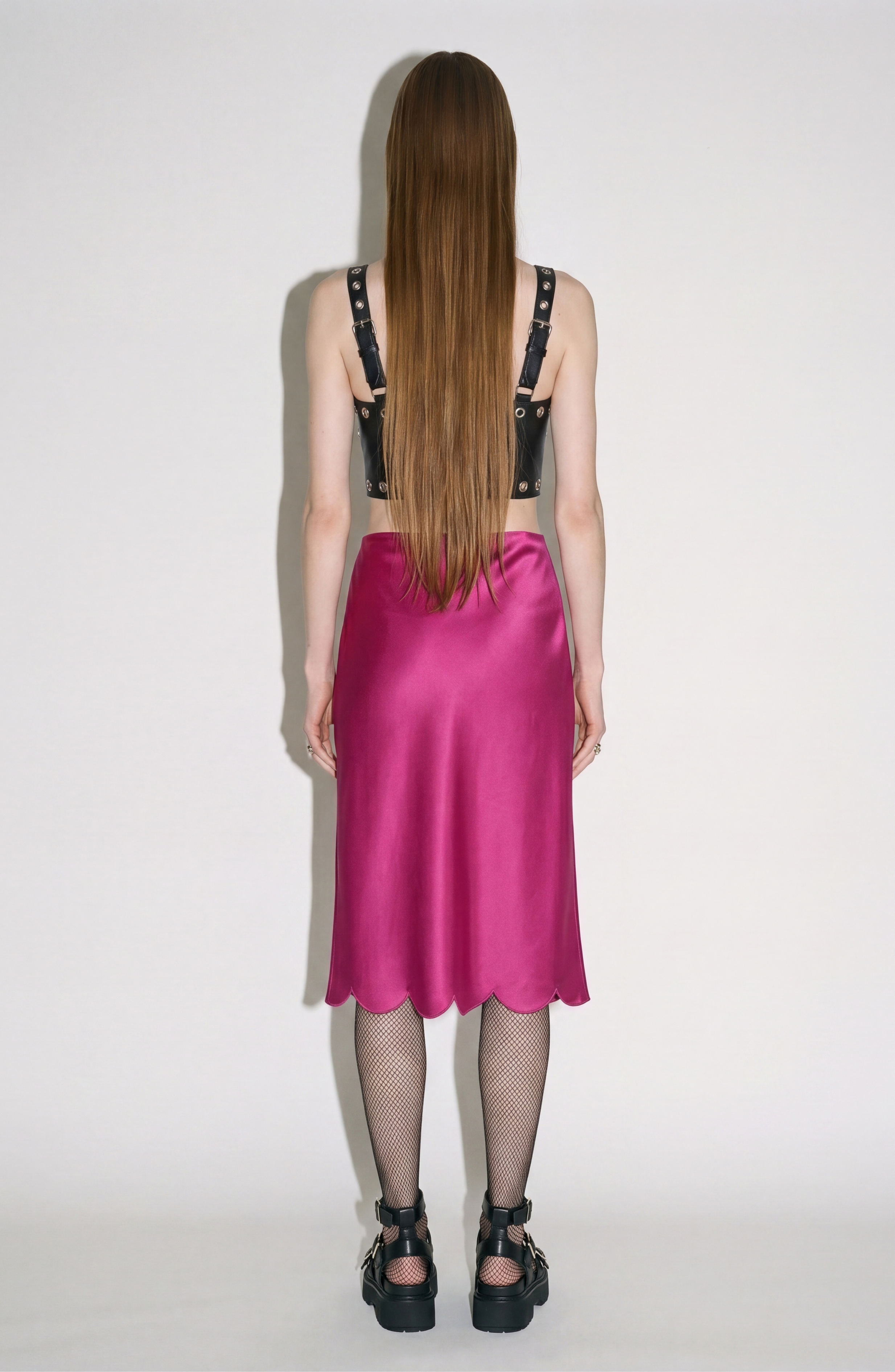 A person with long straight hair stands facing away, wearing the LOULOU Black Leather Top, a shiny pink midi skirt with a scalloped hem, fishnet tights, and chunky black shoes against a plain light background.