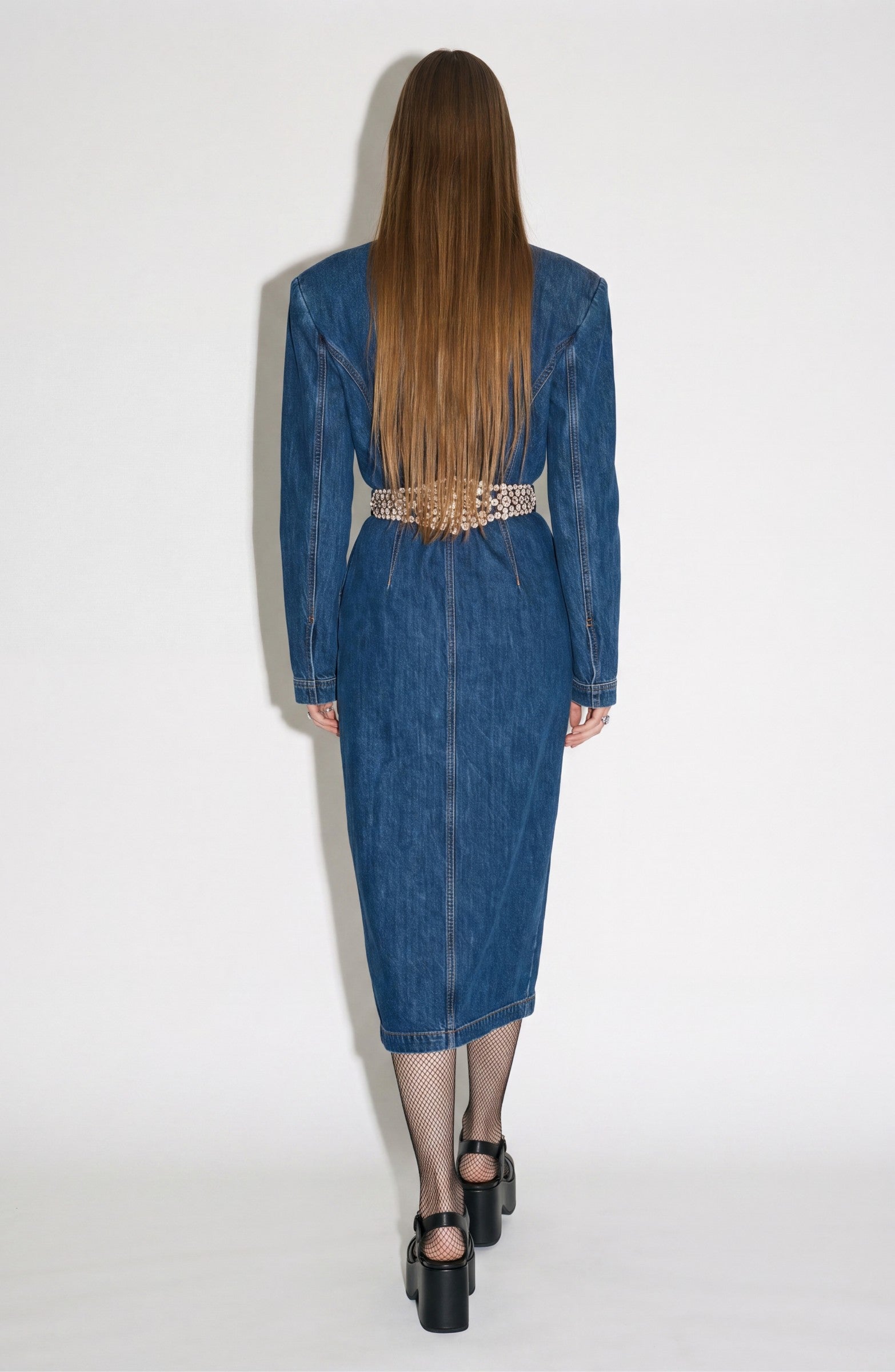 A person with long straight hair stands with their back to the camera against a plain white background, wearing the LOULOU Crystal Belt Denim Dress—a dark indigo midi dress featuring a crystal-embellished belt—paired with fishnet stockings and black platform heels.