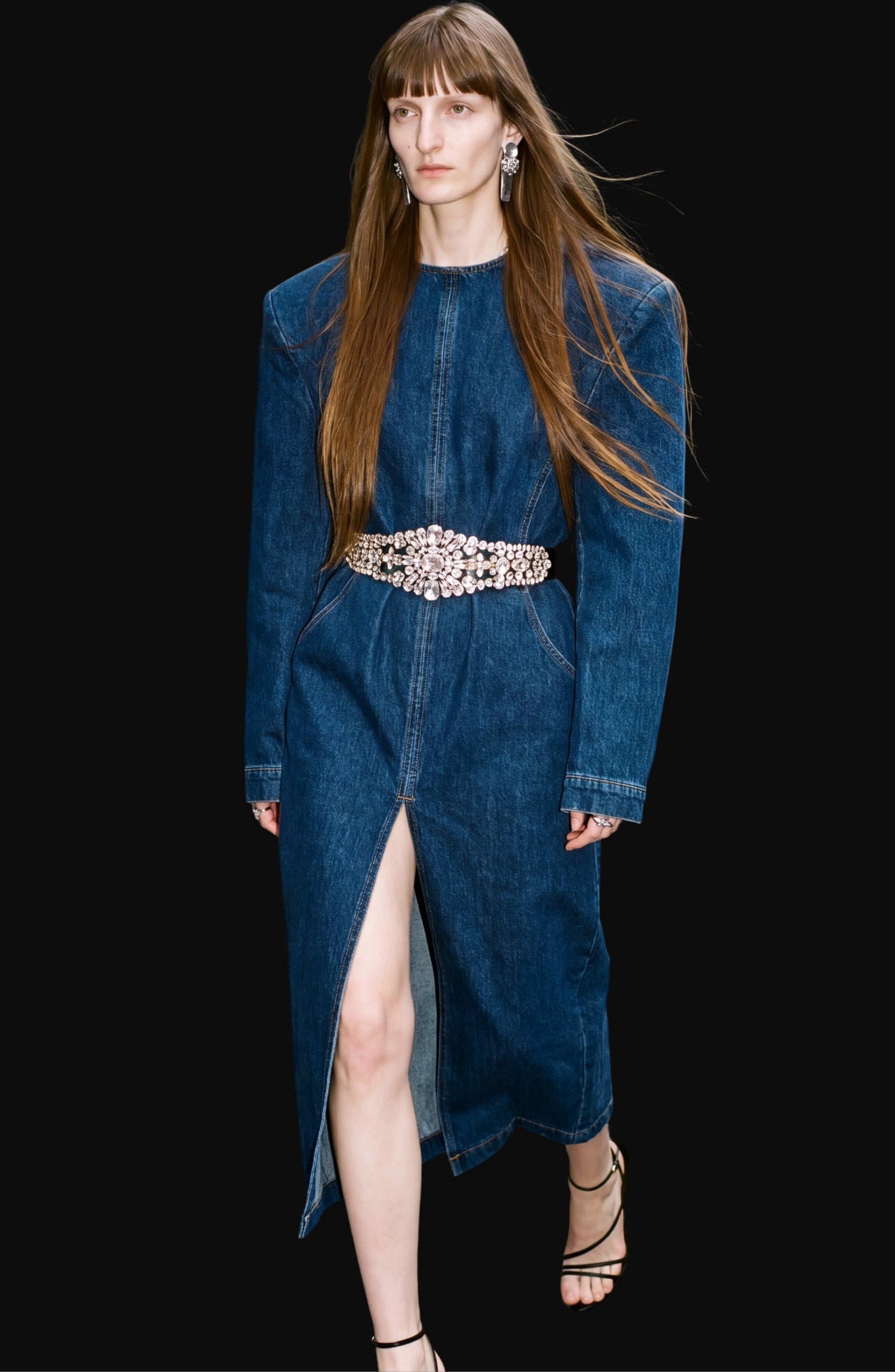 A model showcases LOULOU’s Crystal Belt Denim Dress—a long-sleeve, dark indigo midi dress featuring a front slit and a crystal-embellished belt at the waist—styled with strappy sandals and statement earrings against a black backdrop.