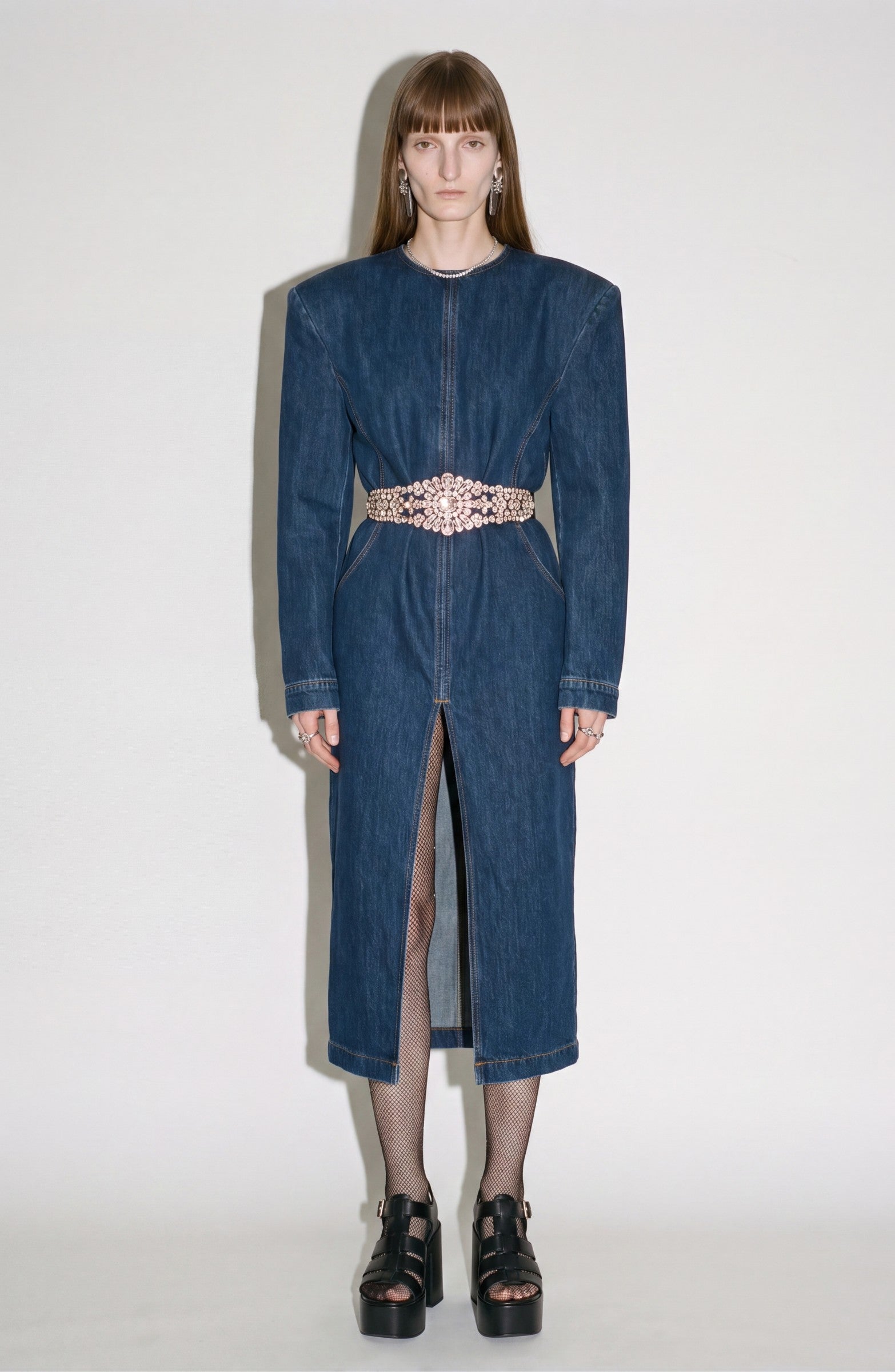 A person stands against a plain background wearing LOULOU’s Crystal Belt Denim Dress—a structured dark indigo midi dress featuring sharp shoulders, a front slit, and a crystal-embellished belt—paired with fishnet tights and black platform sandals.