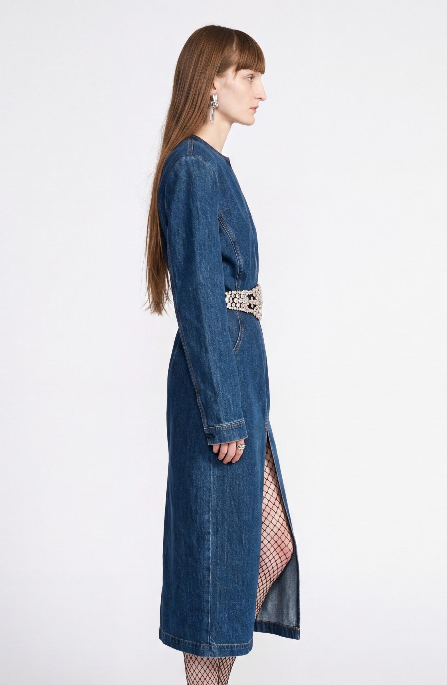 A woman in profile stands against a plain white background, wearing the LOULOU Crystal Belt Denim Dress—a long-sleeve dark indigo midi dress featuring a crystal-embellished belt at the waist—paired with fishnet stockings and statement earrings.