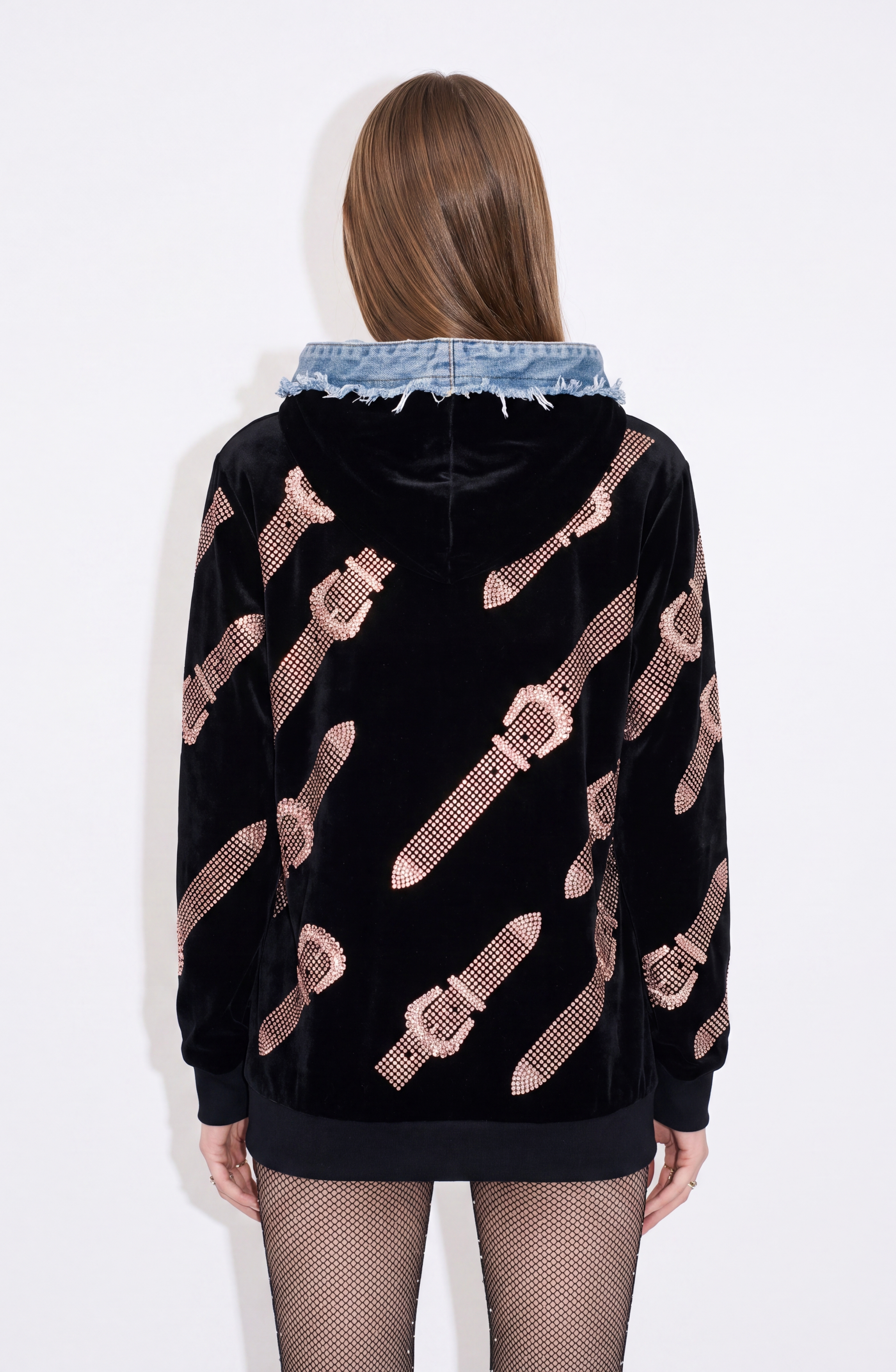 A person with long hair stands facing away from the camera against a plain white background, wearing the LOULOU Crystal Belt Velvet Hoodie in black with a pink belt pattern and a blue denim hood, paired with fishnet tights.