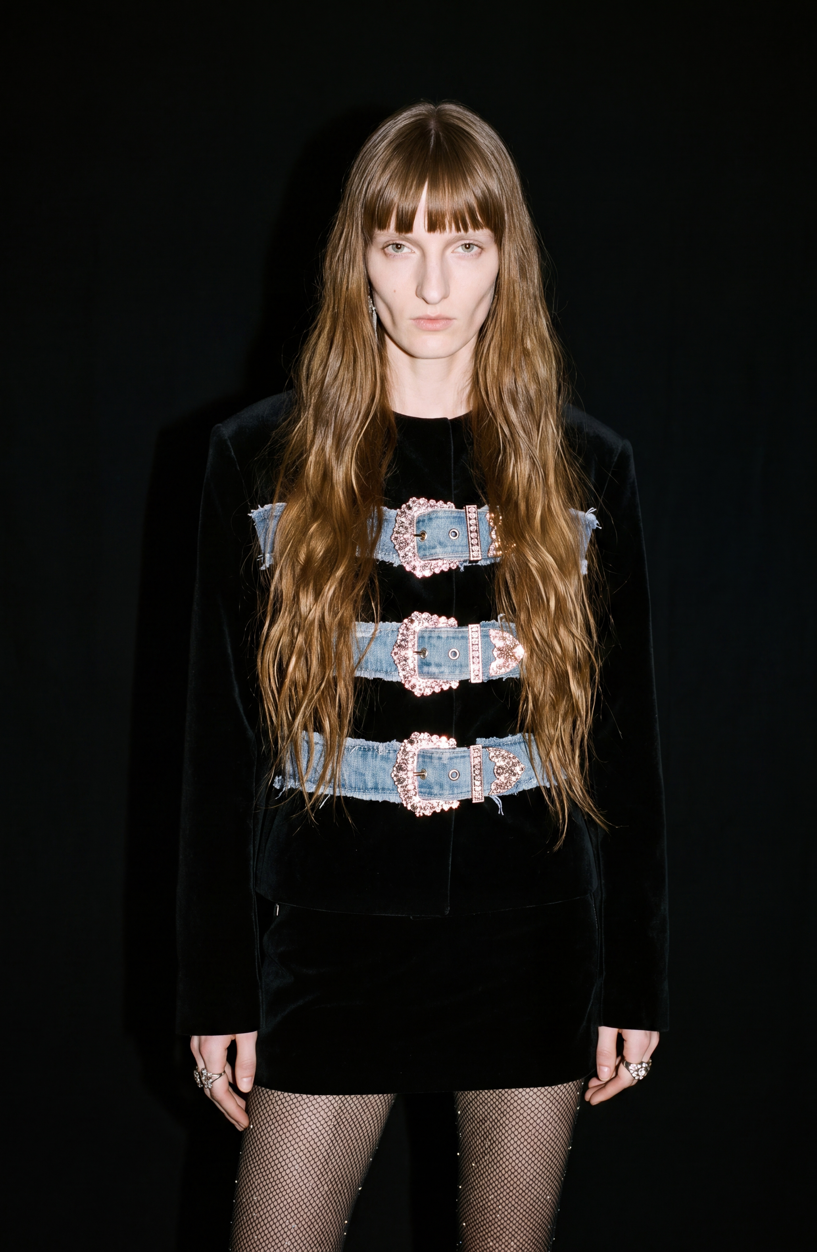 A person with long, wavy brown hair wears the LOULOU Crystal Buckle Velvet Jacket, featuring three blue crystal-embellished buckles, paired with fishnet tights and standing against a black background.