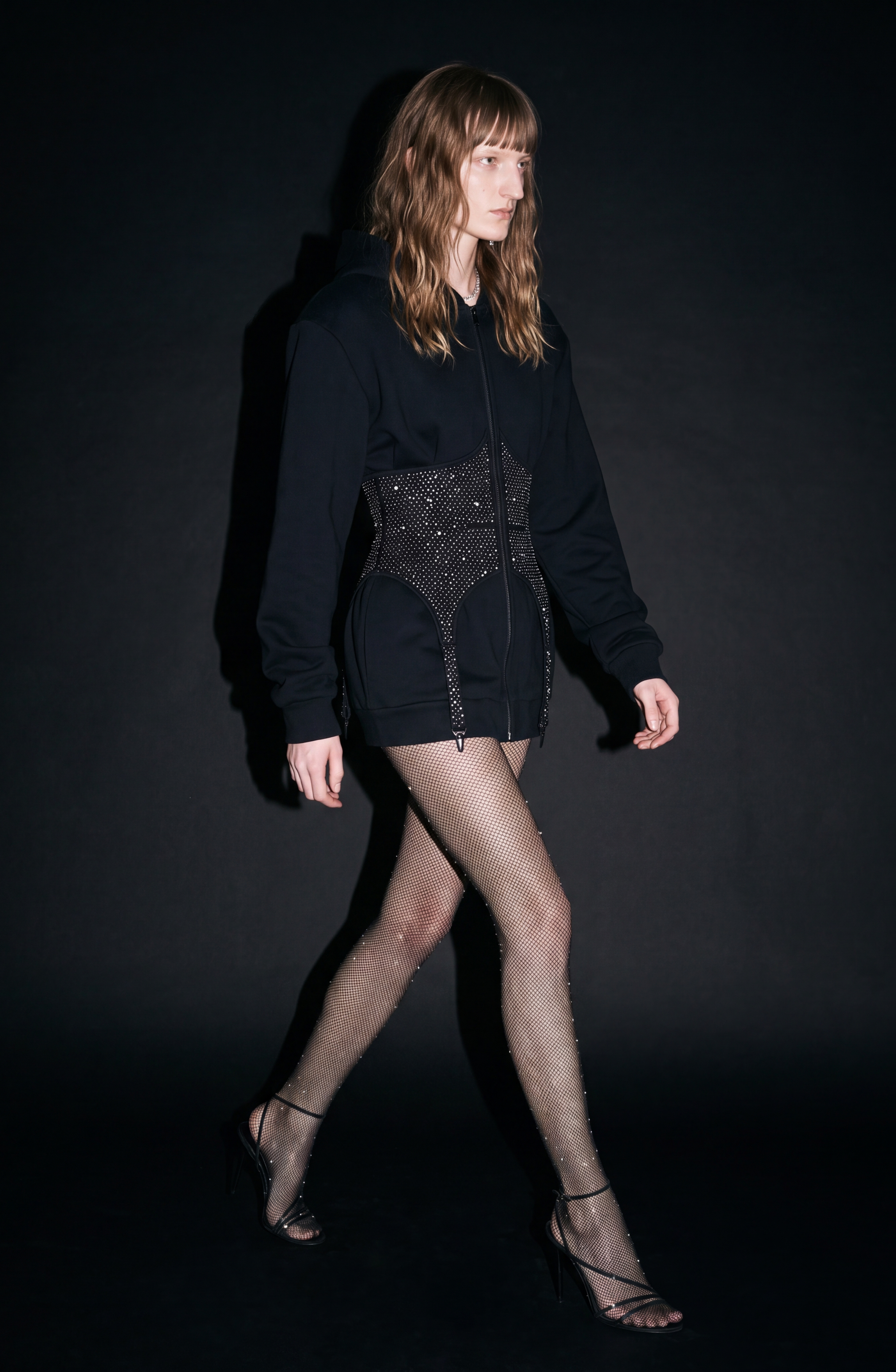 A person with long, wavy hair walks against a dark background, wearing the LOULOU Crystal Corset Zip Hoodie that creates a structured silhouette, styled with sheer rhinestone tights and black high-heeled sandals.