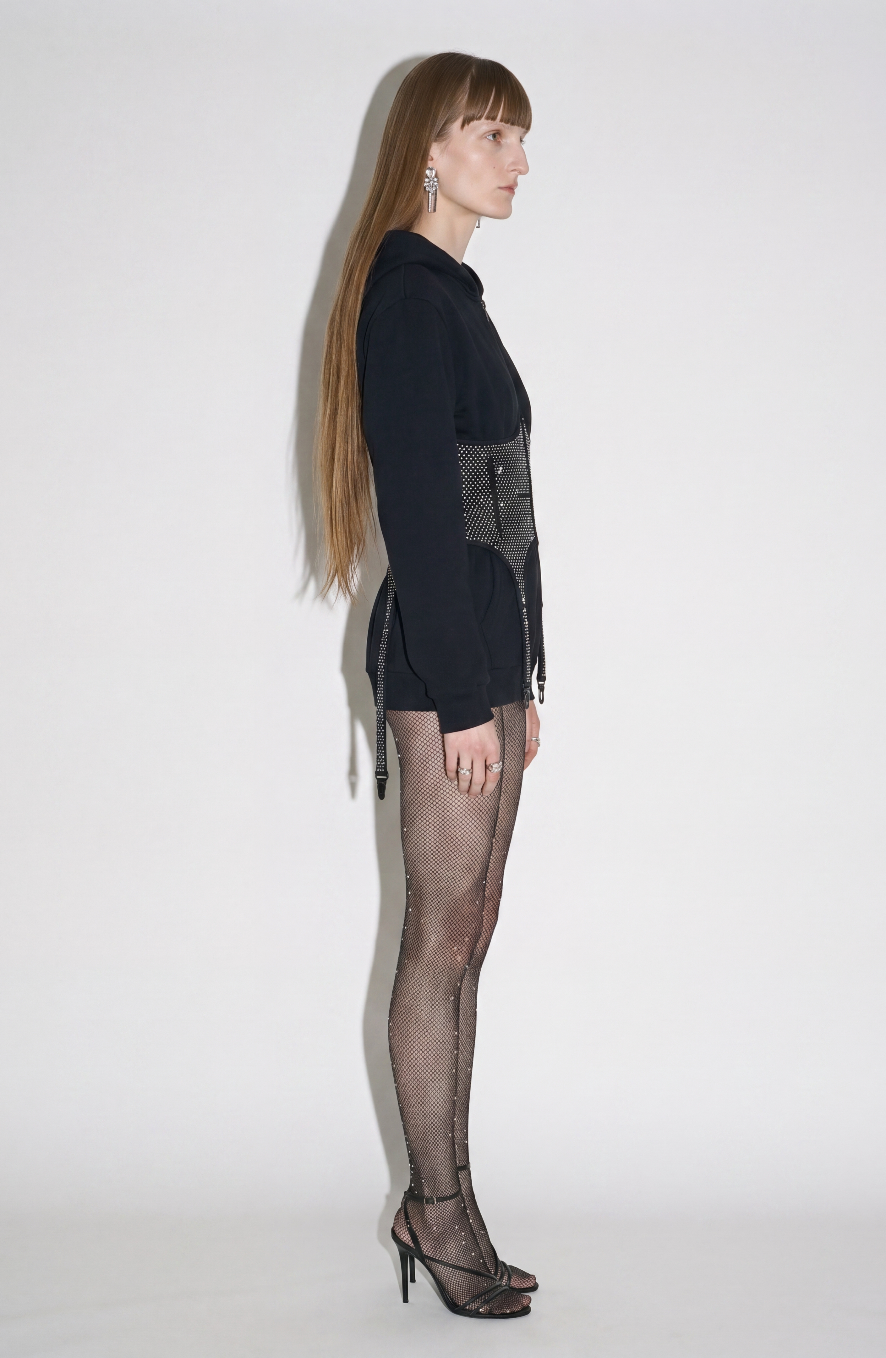 A person with long straight hair stands in profile facing right, wearing the LOULOU Crystal Corset Zip Hoodie, mesh tights, and black high-heeled sandals against a plain white background.