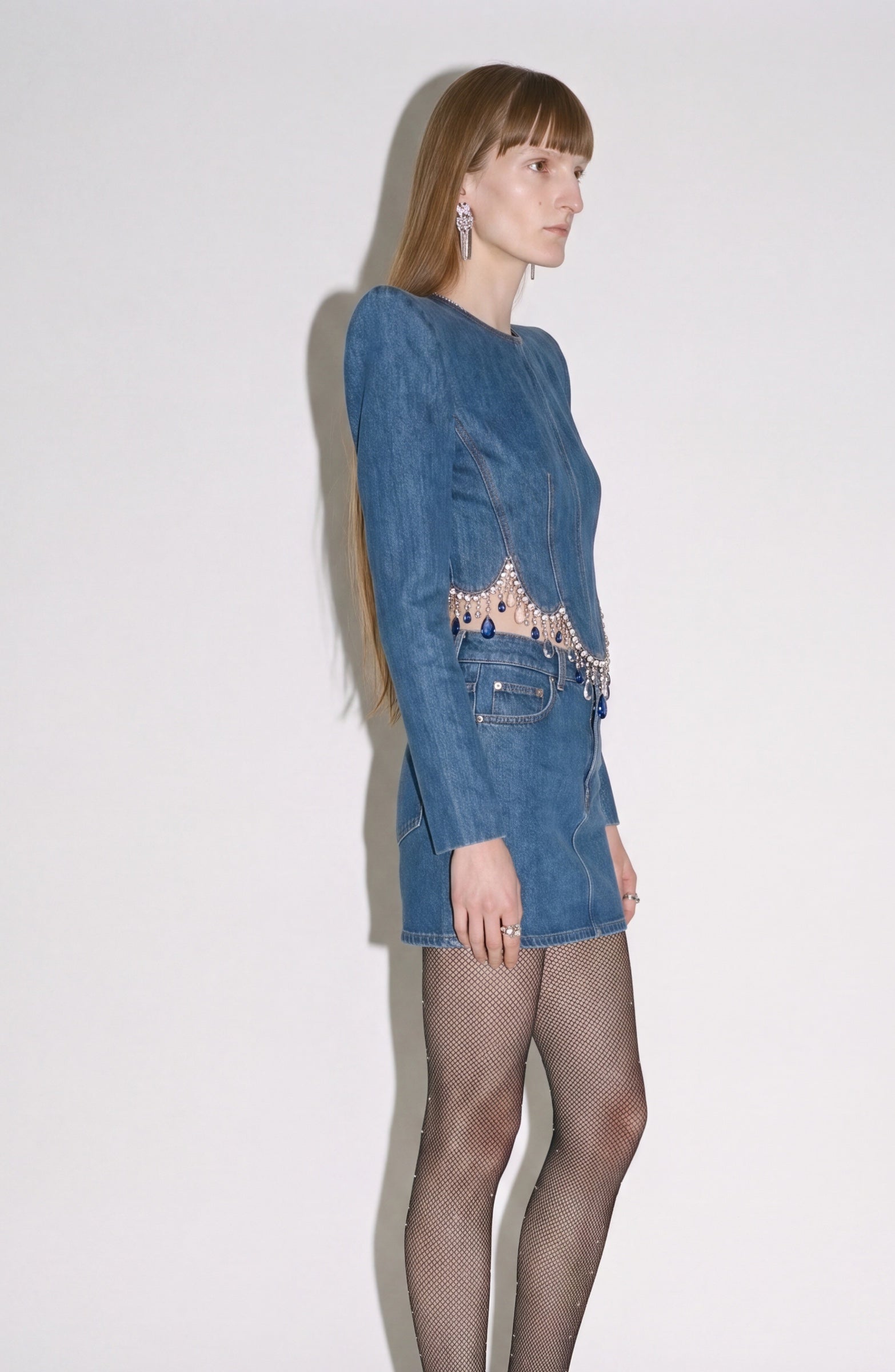 A woman stands sideways wearing the LOULOU Crystal Fringe Denim Top, featuring long sleeves, a beaded waistband, and a subtly sculpted panelled silhouette. She pairs it with fishnet tights and statement earrings. Her long straight hair frames her face as she poses against a plain white background.