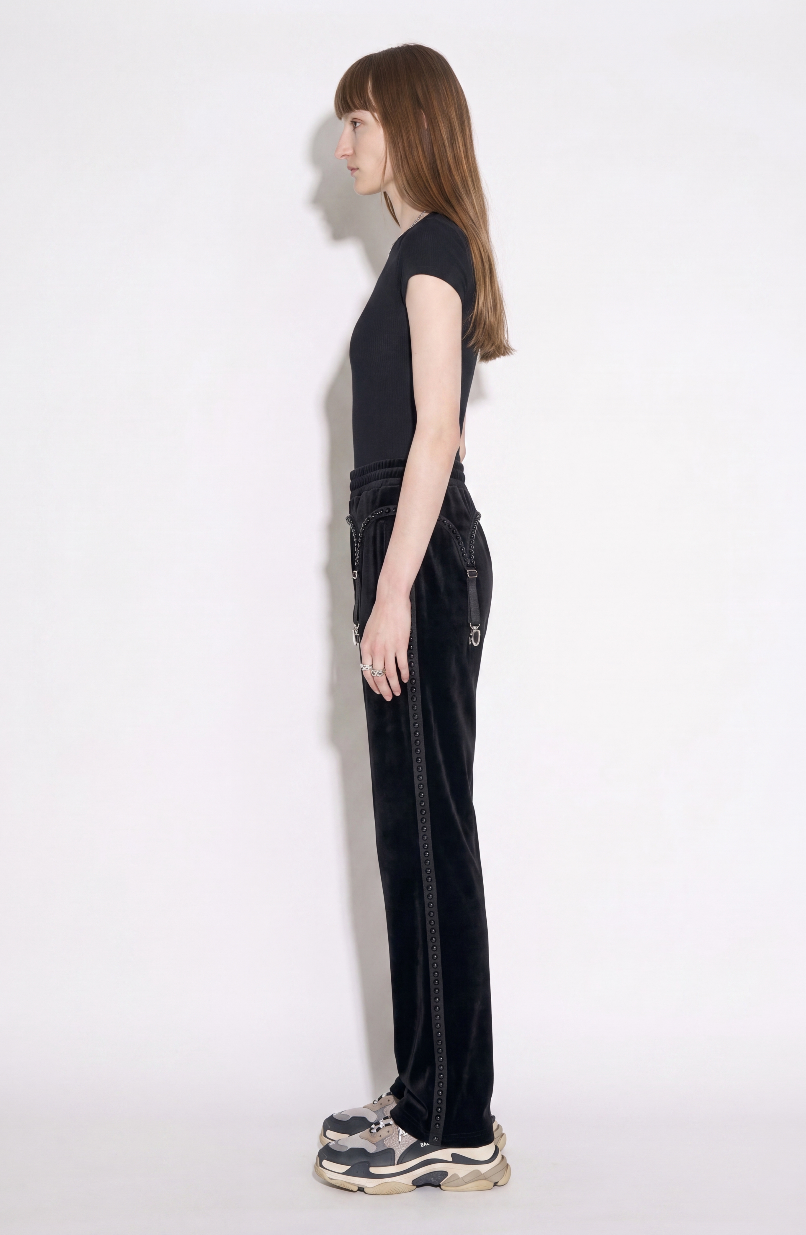 A person with long brown hair stands in profile against a white background, wearing a black t-shirt and LOULOU's Crystal Suspender Velvet Trousers, paired with chunky sneakers.