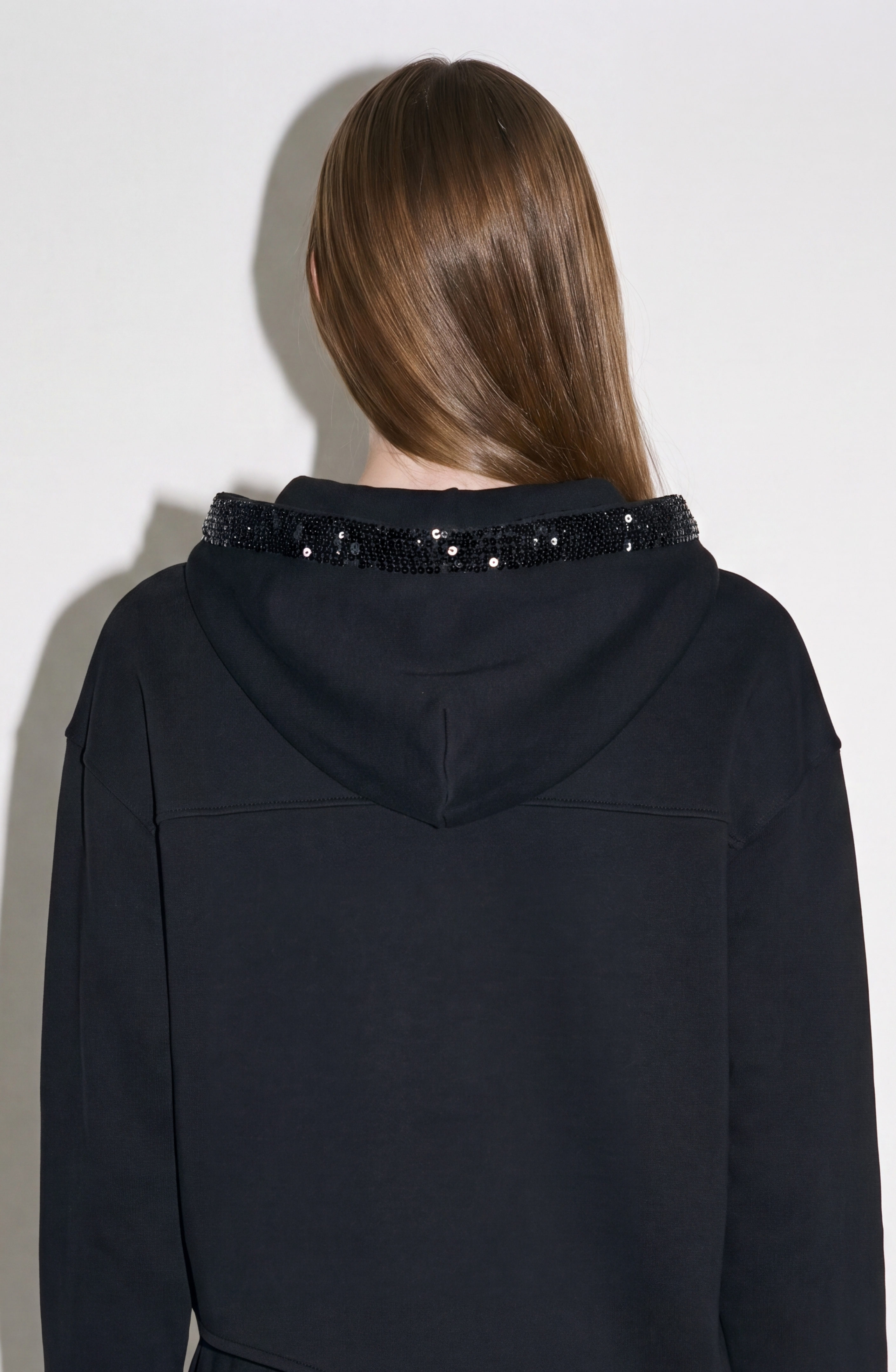 A person with long, straight brown hair is seen from behind wearing LOULOU’s Crystal Trim Asymmetrical Hoodie—an oversized black hoodie made from heavyweight cotton jersey, adorned with crystal embellishments around the edge of the hood—while standing against a plain light-colored wall.