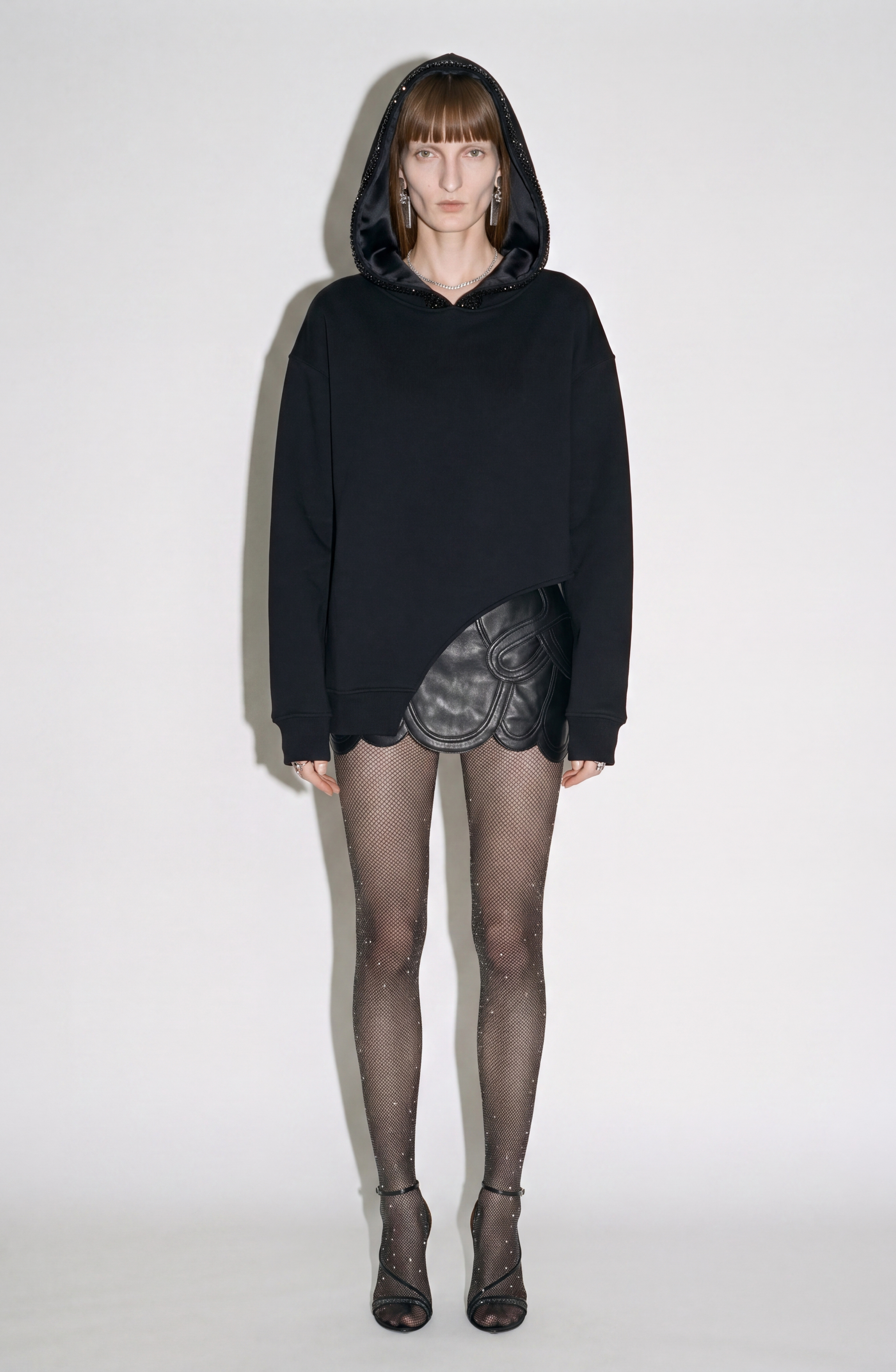 A person stands against a plain background wearing the LOULOU Crystal Trim Asymmetrical Hoodie, an oversized black piece crafted from heavyweight cotton jersey, paired with a black textured skirt, sheer crystal embellished tights, and black open-toe heels.