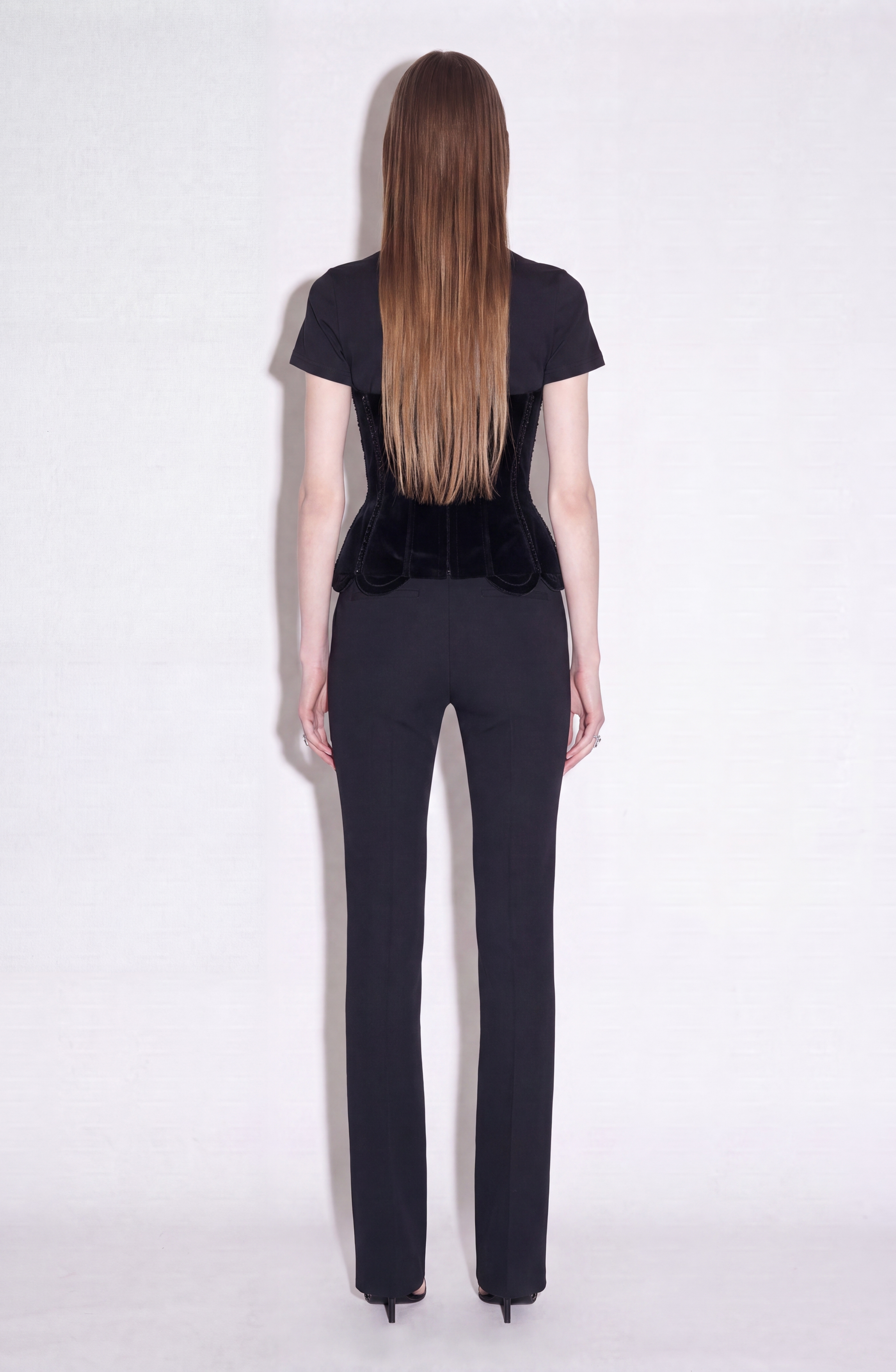 A person with long straight hair, wearing the LOULOU Crystal Velvet Corset T-Shirt, fitted black pants, and black heels stands facing a white background with their back to the camera.