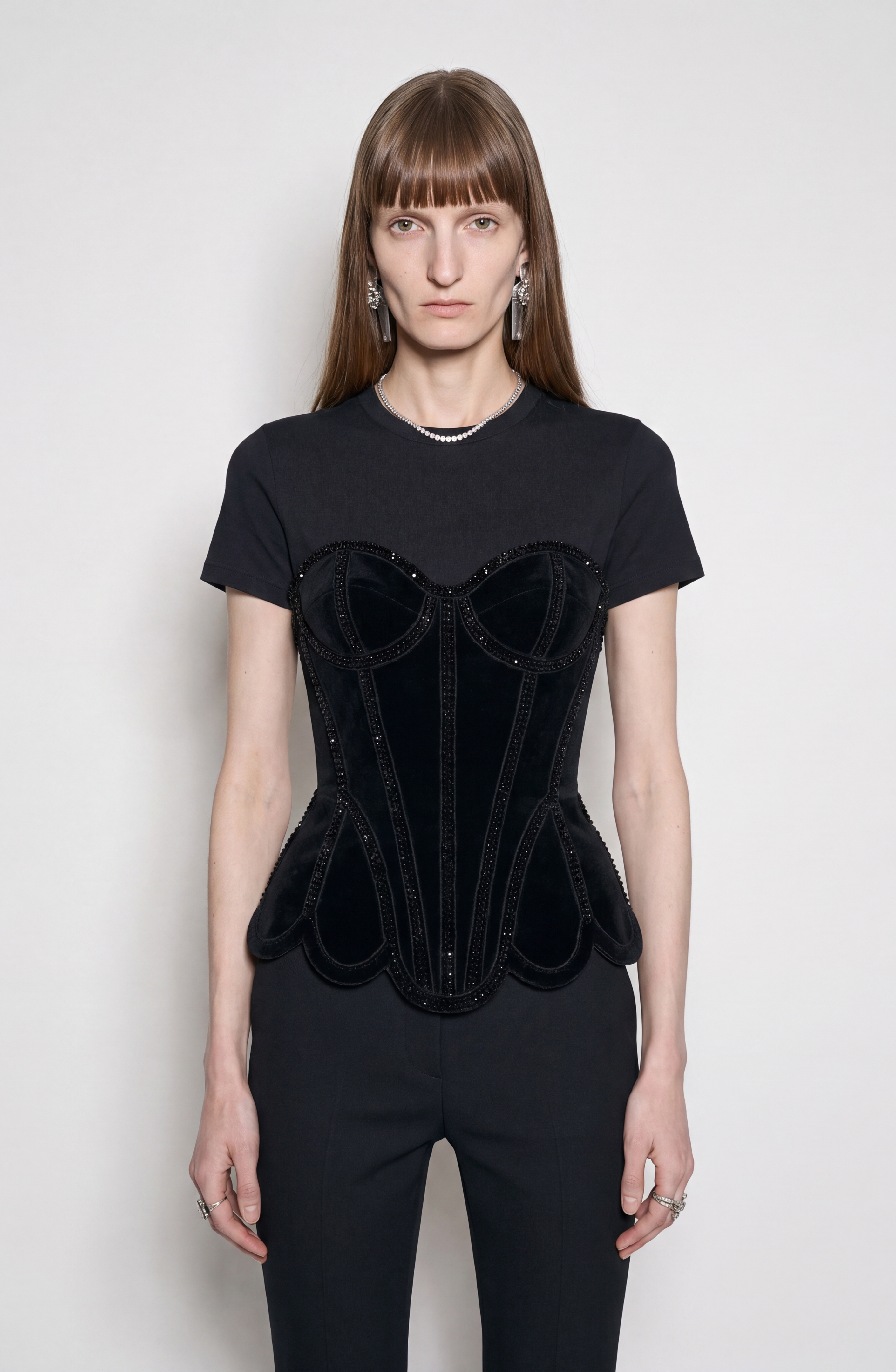 A model with long brown hair and bangs wears the LOULOU Crystal Velvet Corset T-Shirt, featuring a black cotton jersey base and a black velvet corset top detailed with beaded trim. She pairs it with black trousers and accessorizes with a delicate necklace, drop earrings, and silver rings. The photo is set against a plain white background.