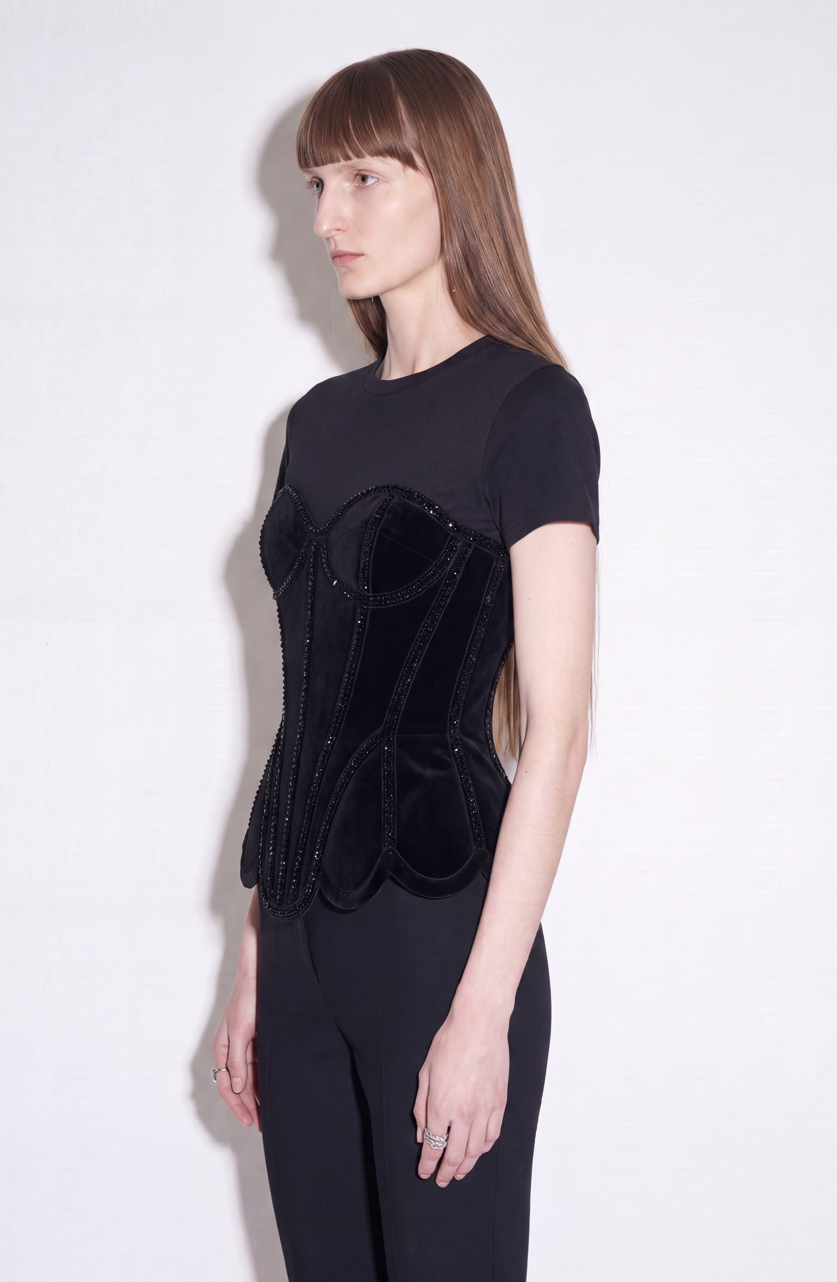 A person with long, straight brown hair is wearing the LOULOU Crystal Velvet Corset T-Shirt—a fitted black cotton jersey top featuring a beaded velvet corset detail—paired with black pants and standing in profile against a plain light background.