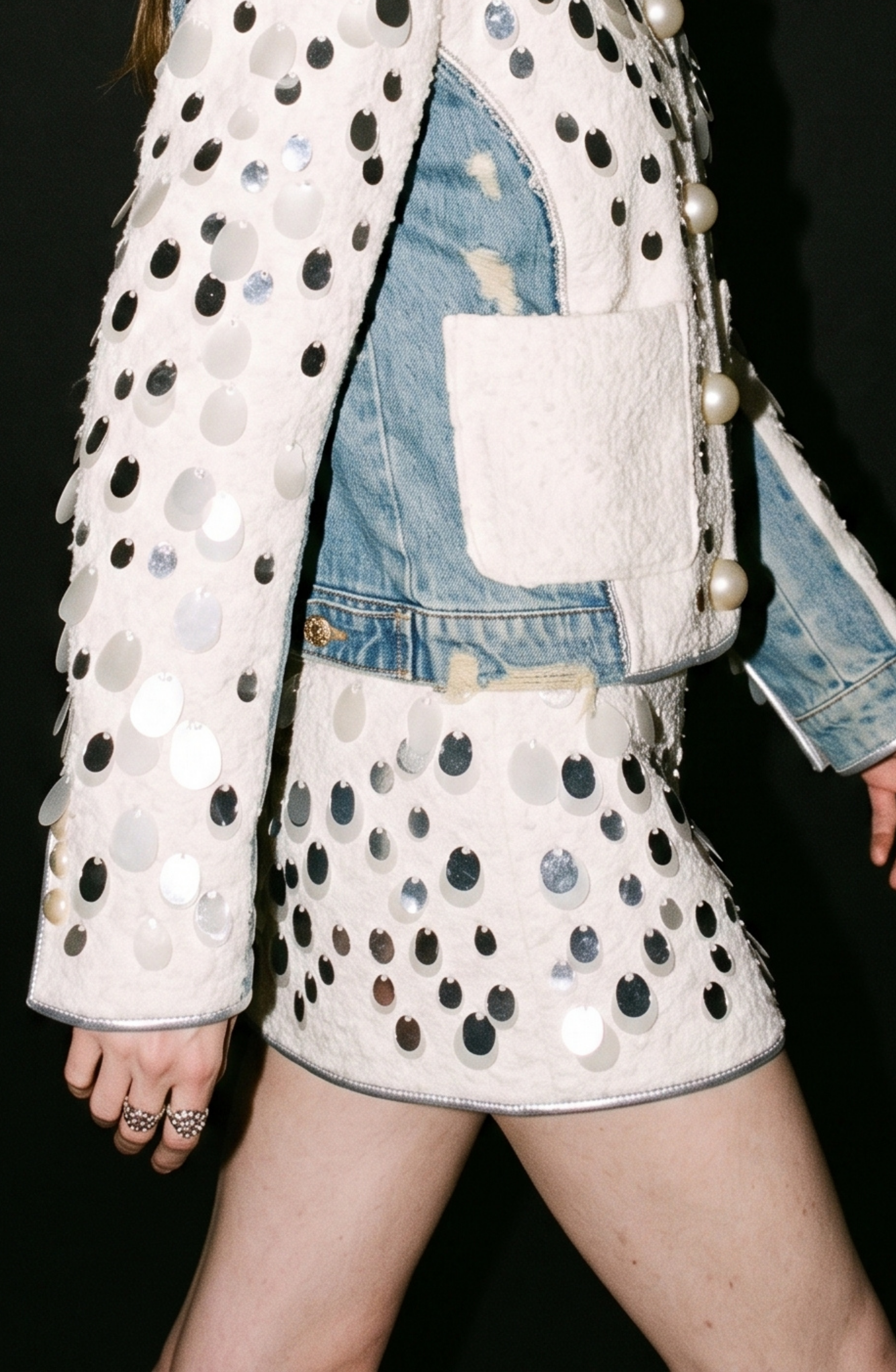 A person wearing the LOULOU Embellished Bouclé Mini Skirt paired with a textured jacket adorned with large silver paillettes over a distressed denim jacket featuring a visible denim waistband, standing against a black background.