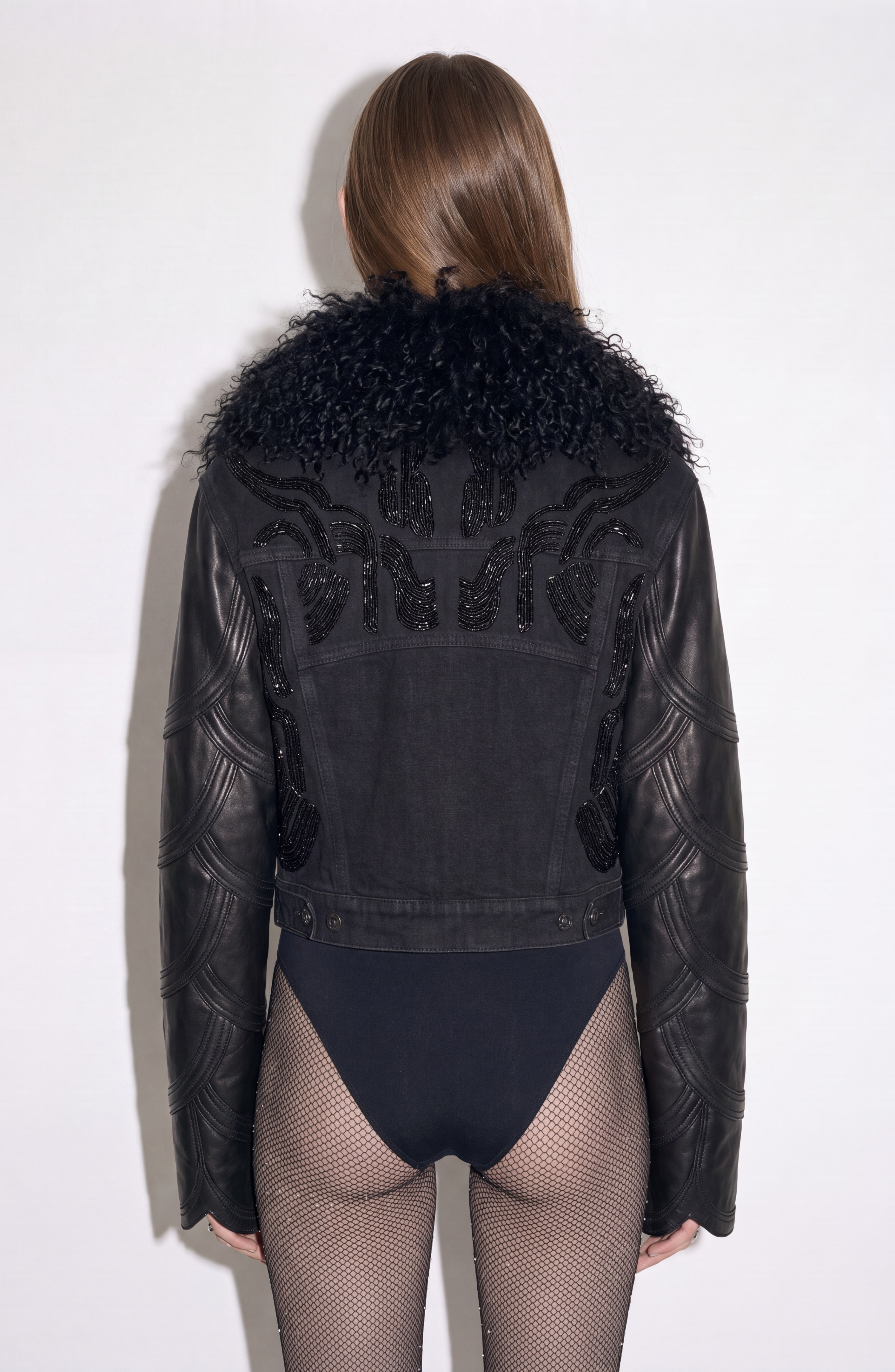 A person wearing the LOULOU Embellished Denim Leather Jacket, featuring quilted leather sleeves and a textured shearling collar, paired with a black bodysuit and fishnet stockings, shown from the back against a plain light background.