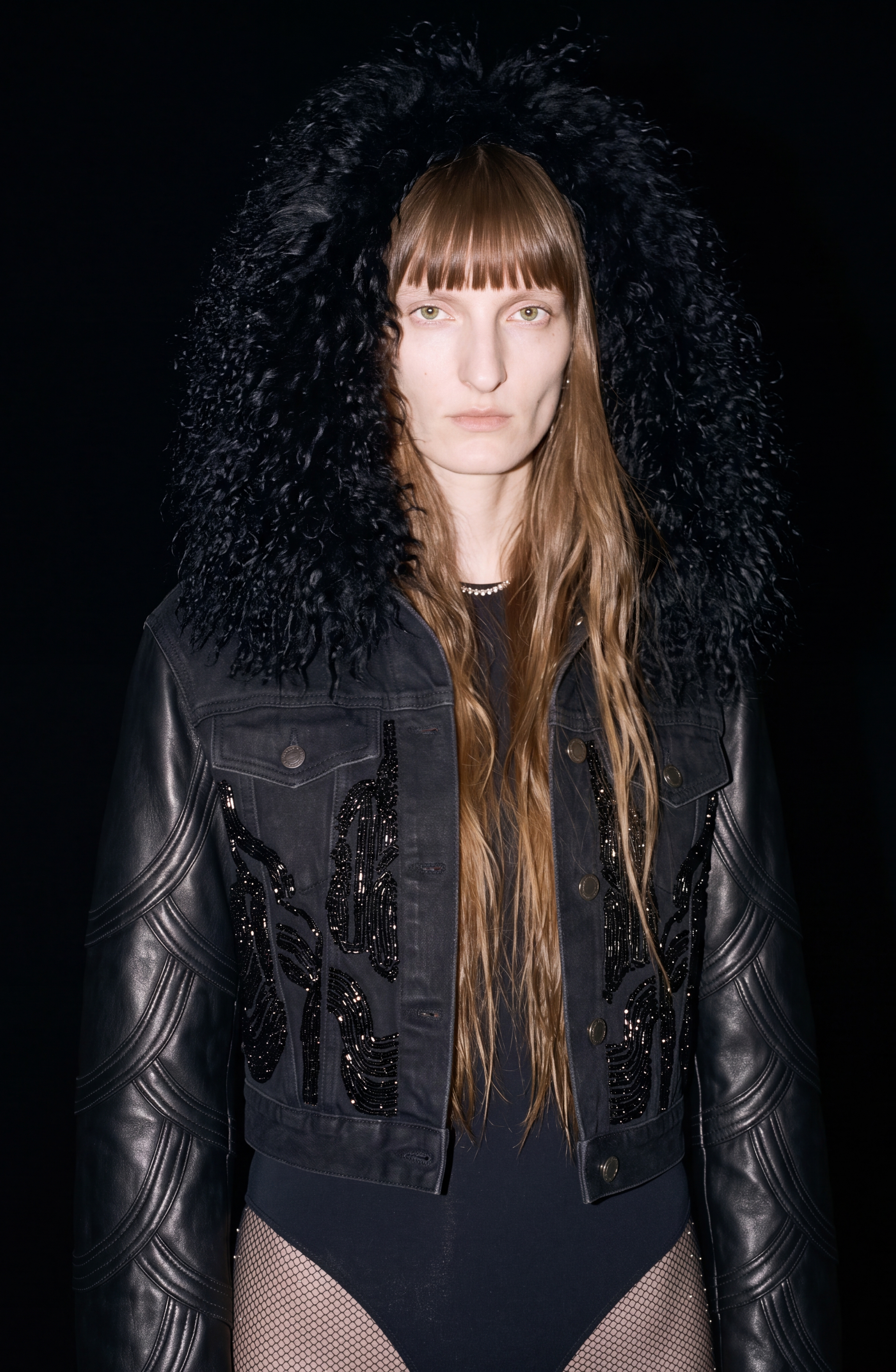 A person with long hair and blunt bangs wears a black faux fur hood and the LOULOU Embellished Denim Leather Jacket, featuring quilted leather sleeves and a shearling collar, paired with mesh tights, standing against a black background.