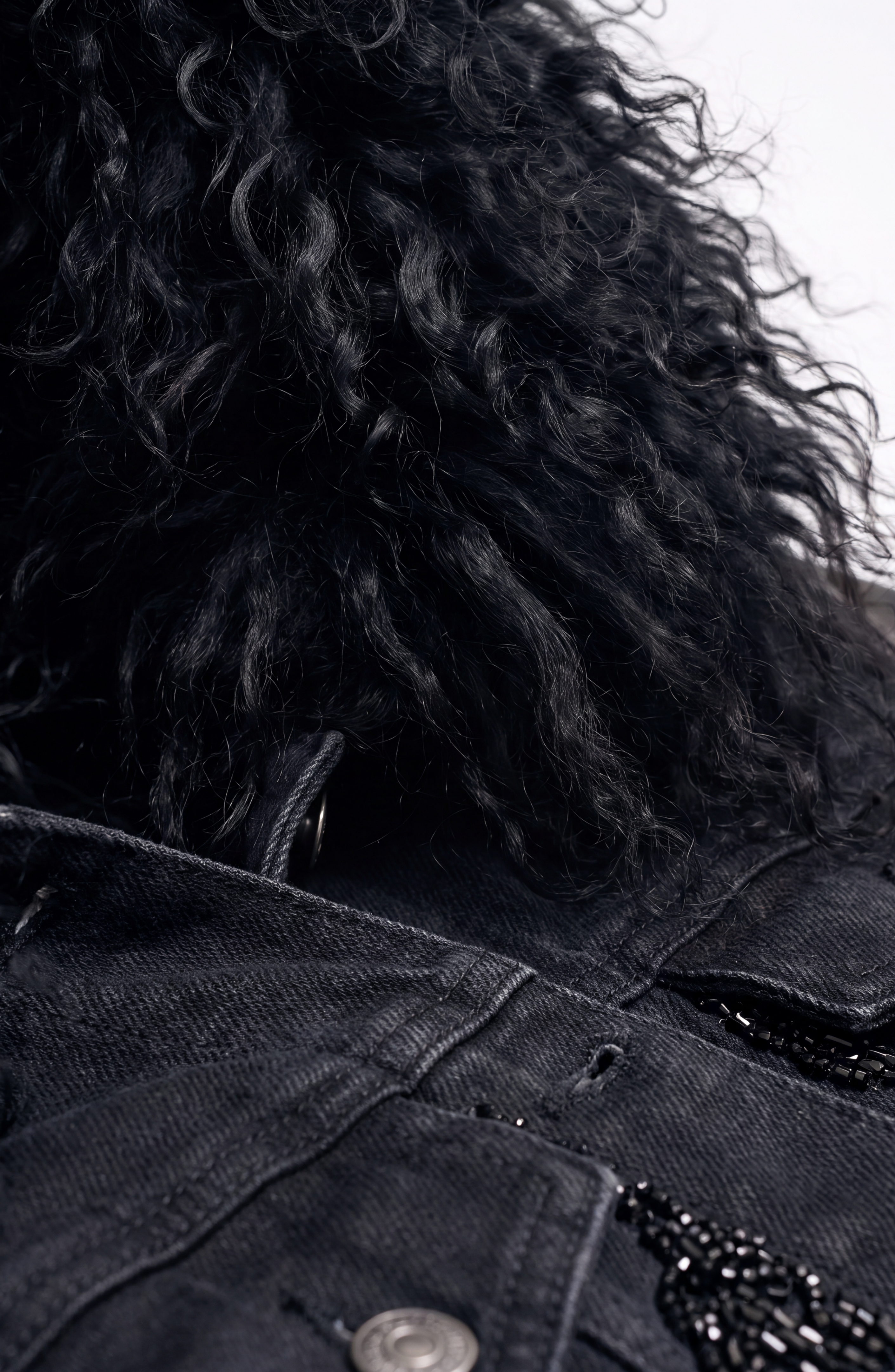 Close-up of curly black hair above the LOULOU Embellished Denim Leather Jacket, featuring a metallic button and decorative black beading. The blurred background draws attention to the rich textures of the hair and the jacket.