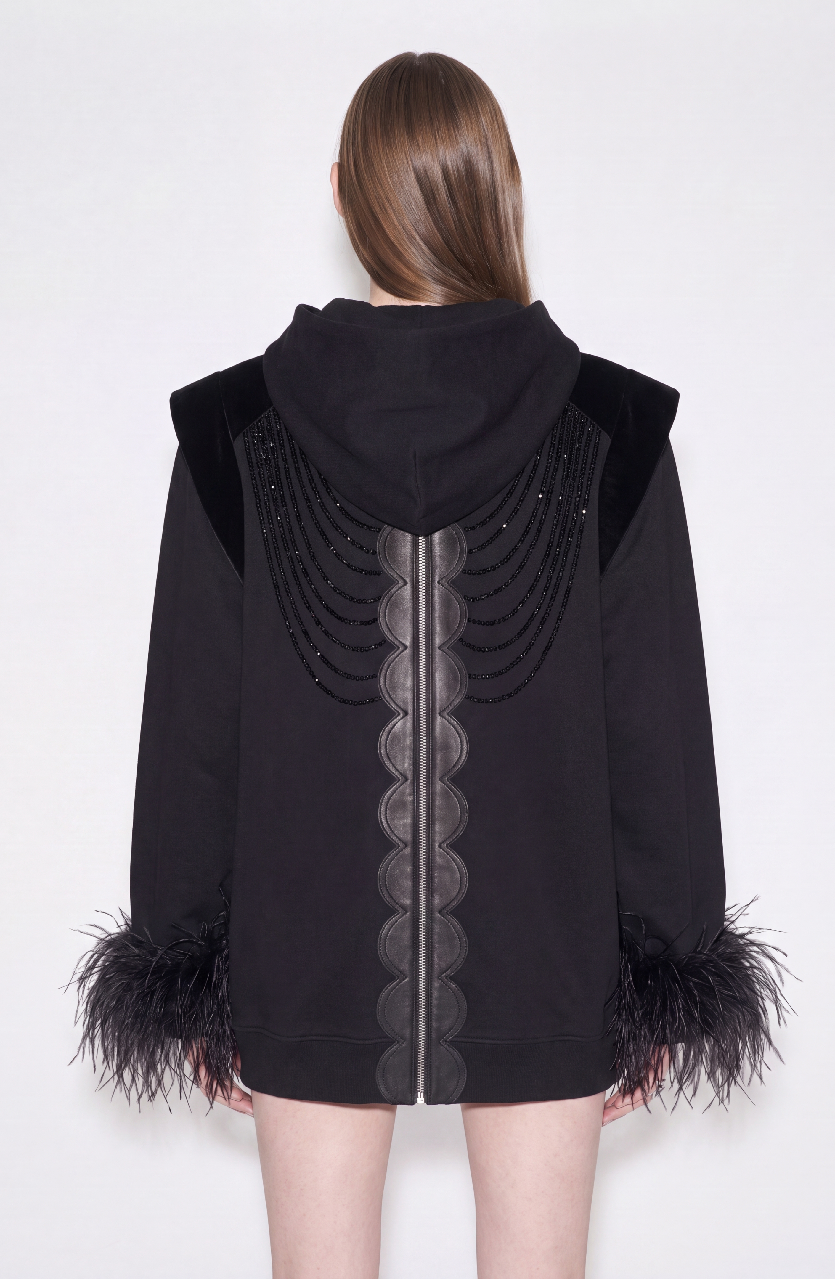 A person stands facing away, wearing the LOULOU Embellished Feather Trim Zip Hoodie in jet black, featuring decorative stitching, velvet accents, and statement feather-trimmed cuffs. Their hair is long and straight, set against a plain white background.