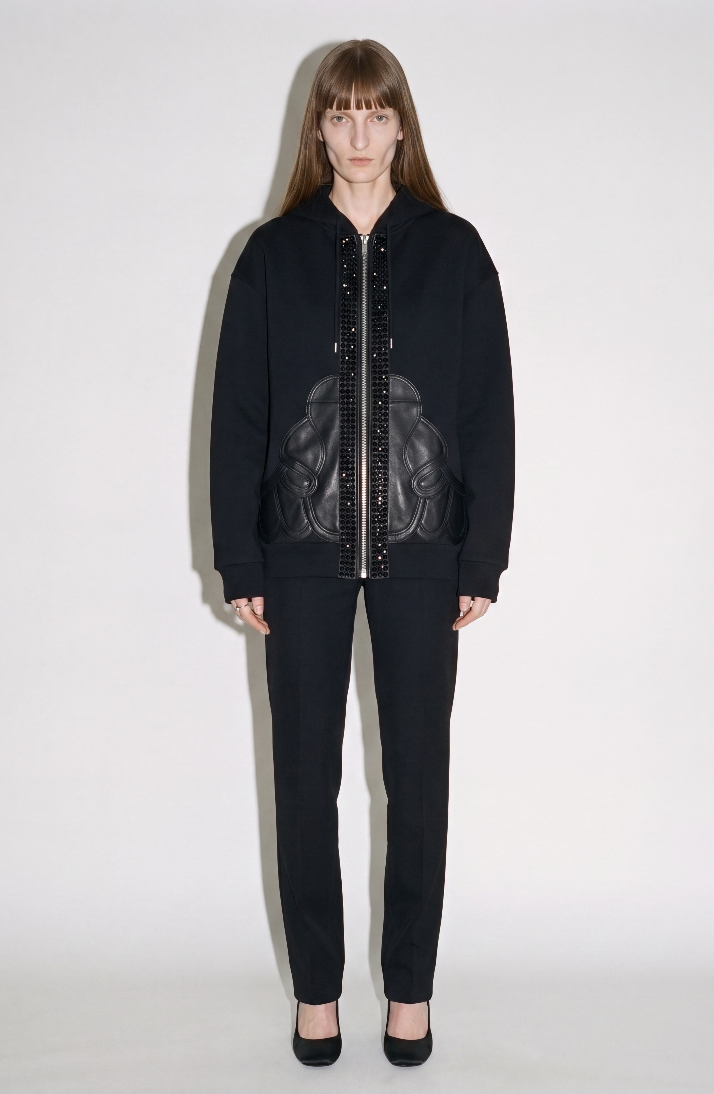 A person with straight, light brown hair wears the LOULOU Embellished Leather Panel Zip Hoodie, featuring decorative patterns and black crystal embellishments, paired with black pants and black heels, standing against a plain white background.