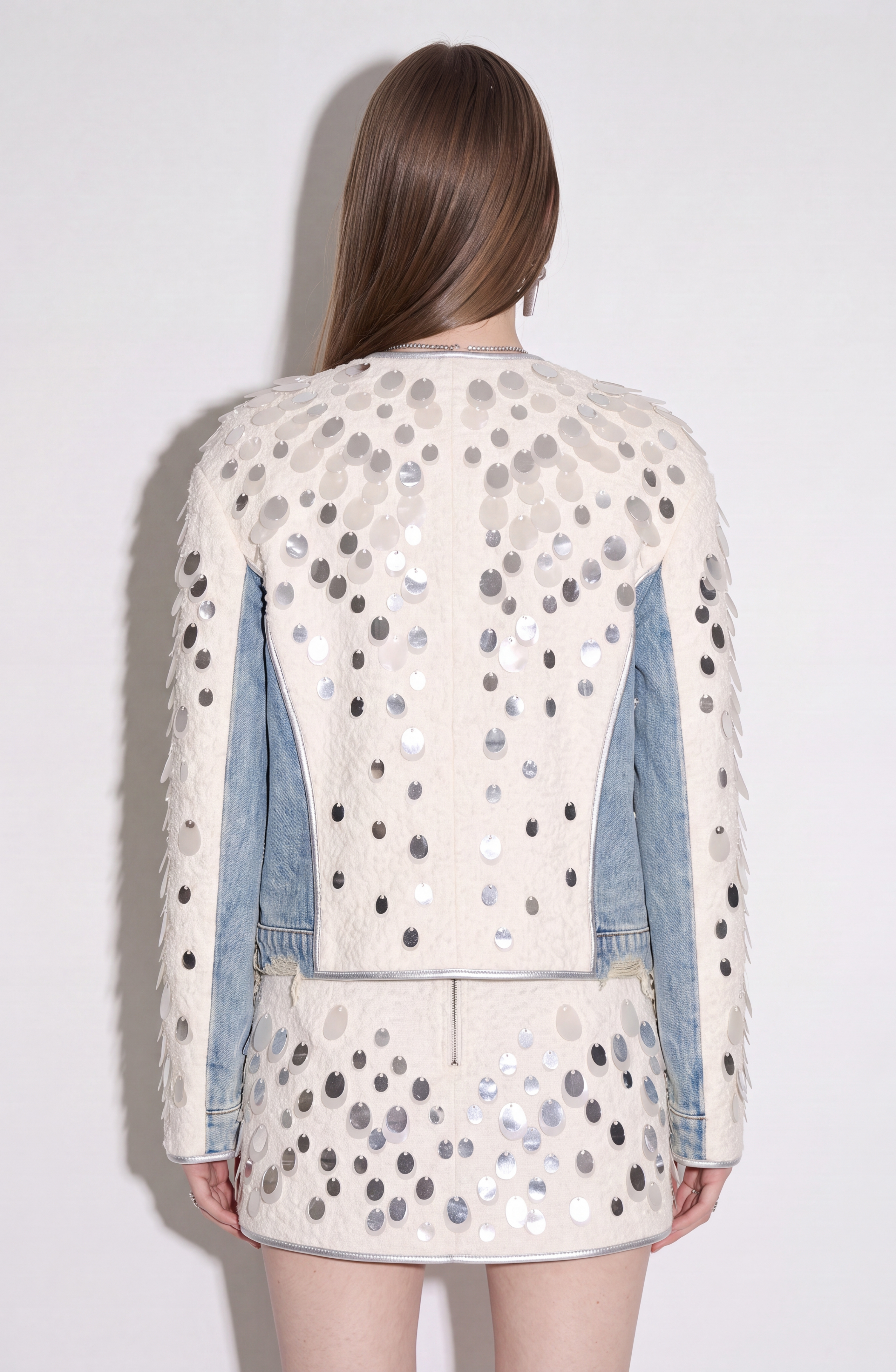 A woman stands with her back to the camera against a plain light background, her straight brown hair down. She wears LOULOU’s Embellished Panelled Cropped Jacket in cream, paired with a matching skirt. The collarless ensemble features large silver sequin embellishments, paneled details with light denim accents, and pearl buttons for added elegance.