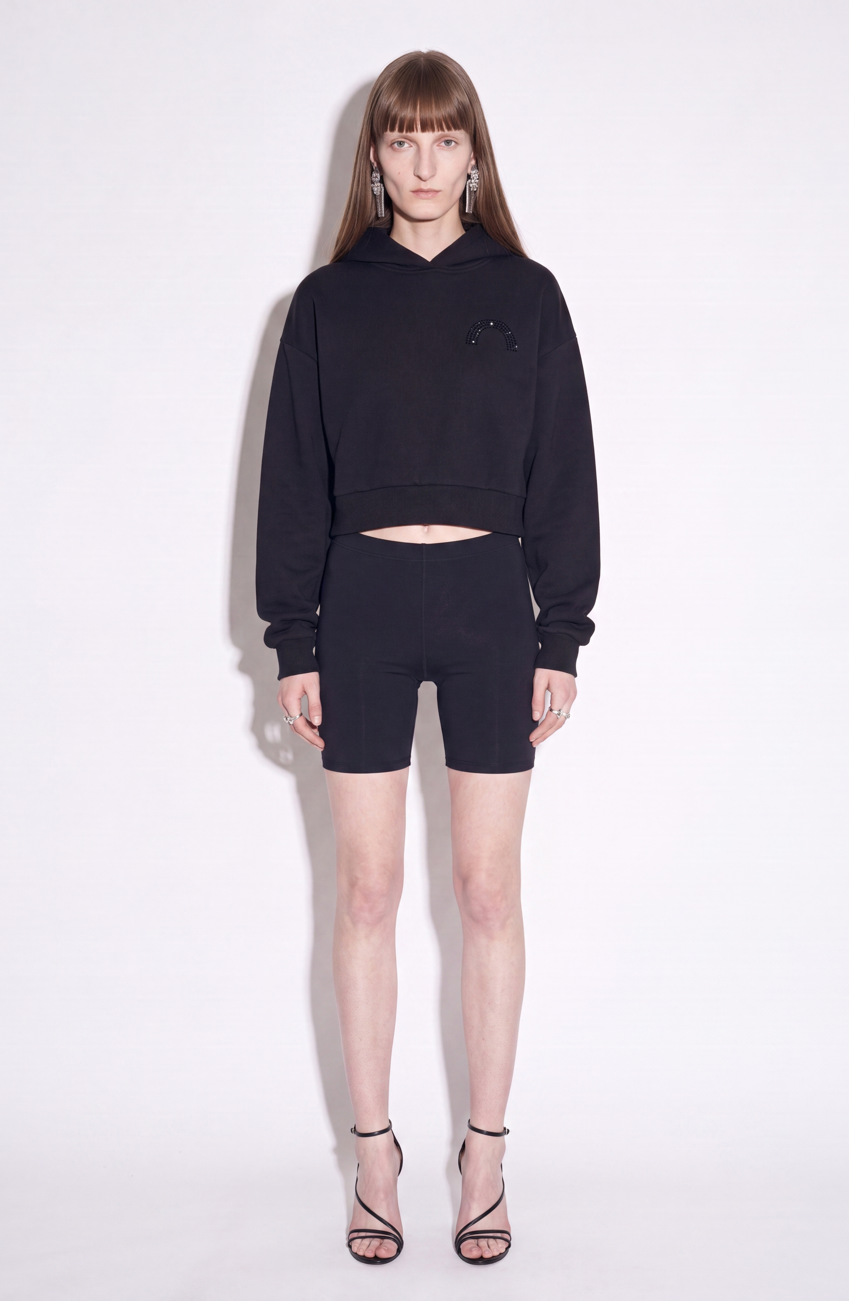 A model with long brown hair stands facing forward against a simple white background, wearing the LOULOU Embellished Tulle Back Cropped Hoodie—a black cotton hoodie featuring a small sparkling rainbow sequin embroidery on the chest—paired with matching black bike shorts and black strappy high-heeled sandals.