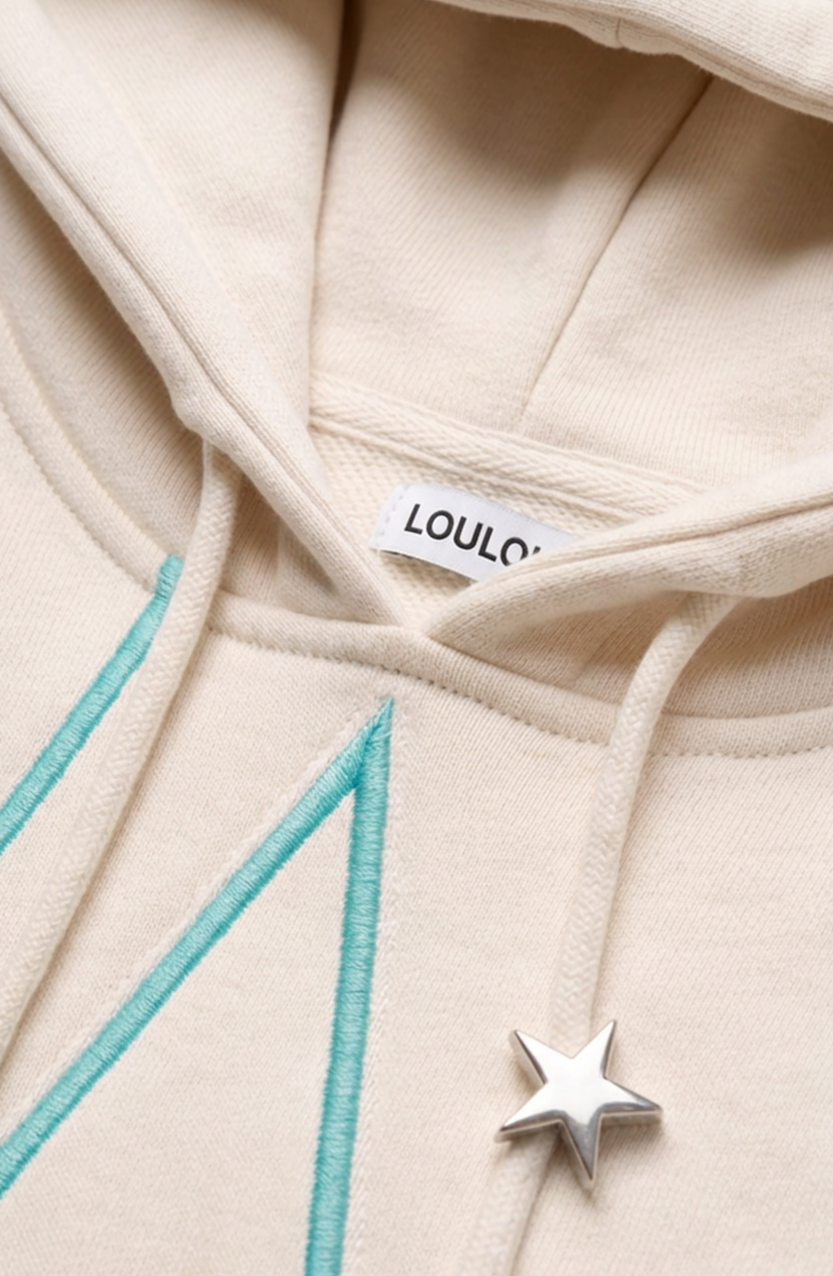 Close-up of the LOULOU Embroidered Star Cotton Hoodie, featuring a cream-colored cotton jersey material, an embroidered turquoise letter, a turquoise star-shaped metal pin on the drawstring, and a visible “LOULOU” tag inside the collar.