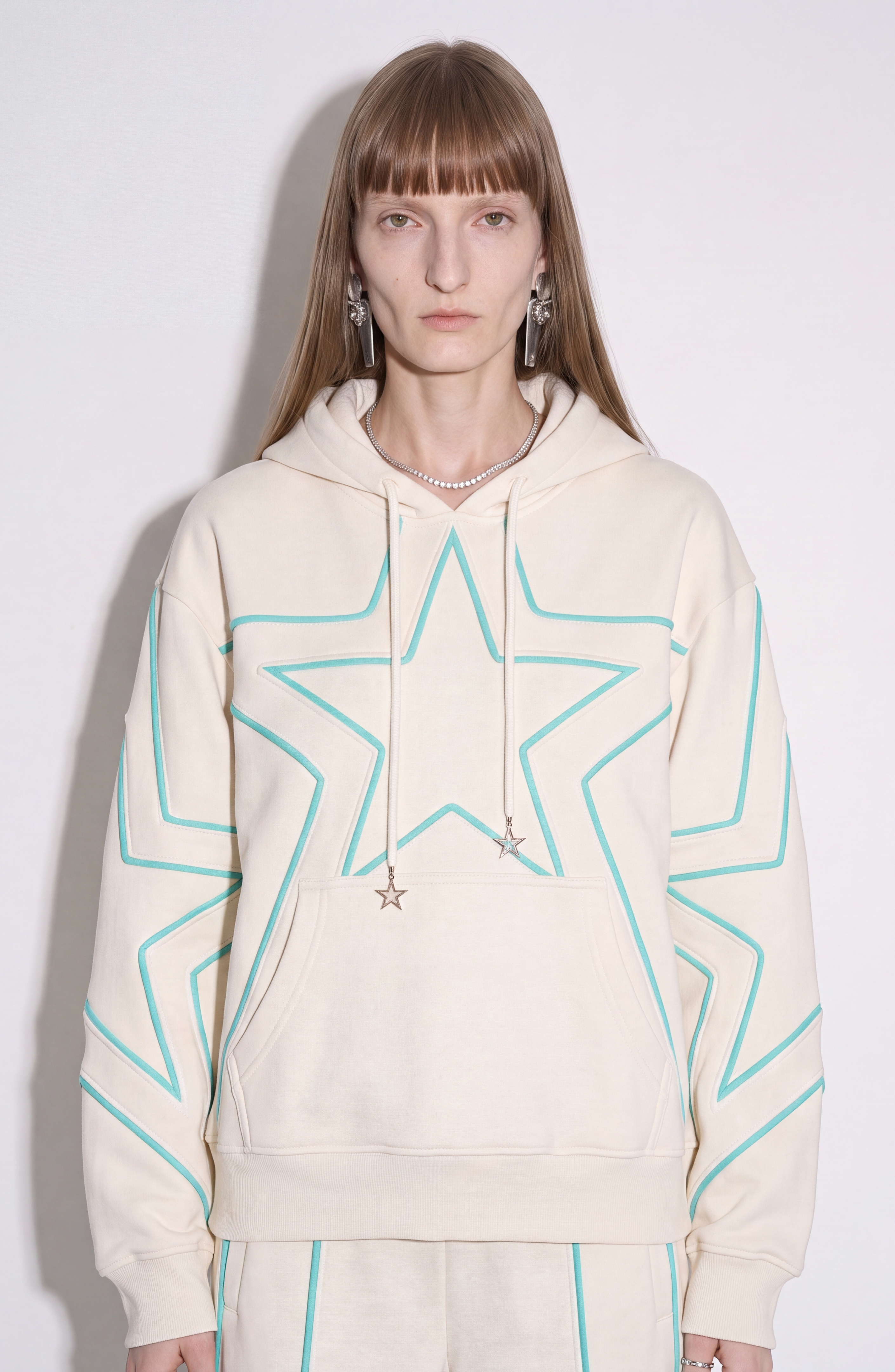 A person with straight, light brown hair wears the LOULOU Embroidered Star Cotton Hoodie in cream—featuring turquoise star outlines—paired with matching pants, star earrings, and a pearl necklace, standing against a plain white background.