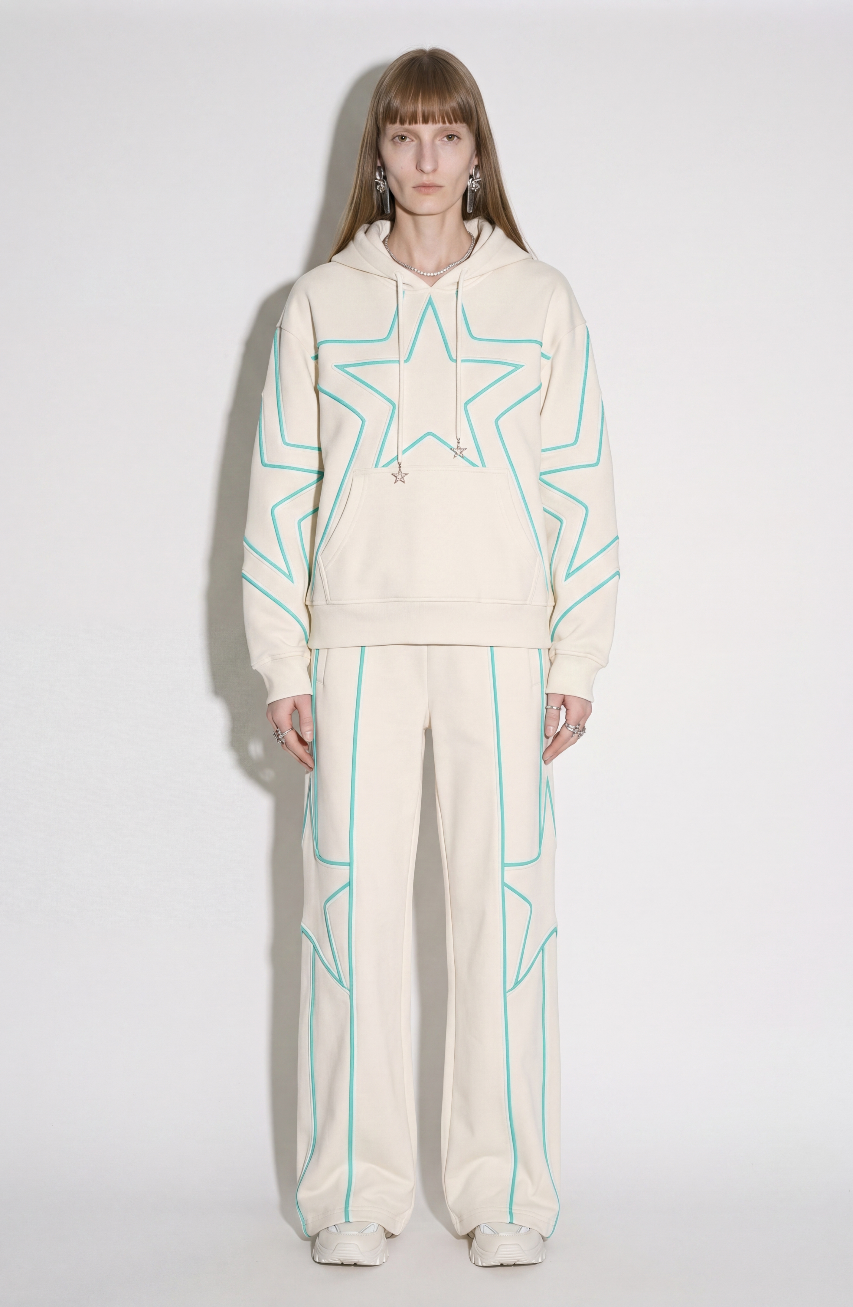 A person stands against a white background wearing the LOULOU Embroidered Star Sweatpants in cream cotton jersey, featuring turquoise star embroidery, paired with a matching hoodie, white sneakers, and statement earrings.