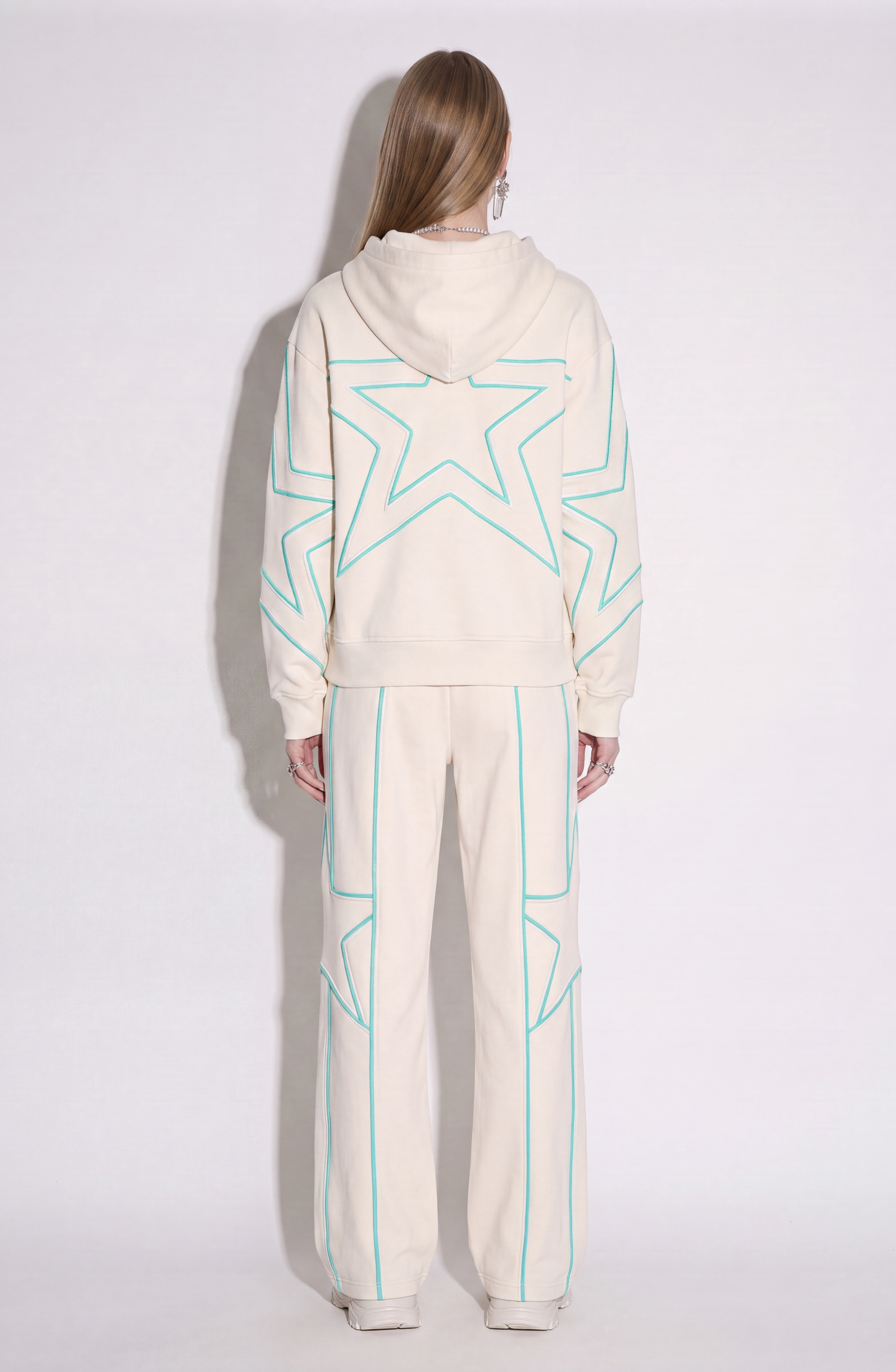 A person stands with their back to the camera, dressed in LOULOU's cream cotton jersey hoodie and Embroidered Star Sweatpants. The wide-leg sweatpants feature outlined teal and turquoise star embroidery. Their long hair is worn down, accessorized with earrings and rings, against a plain neutral background.