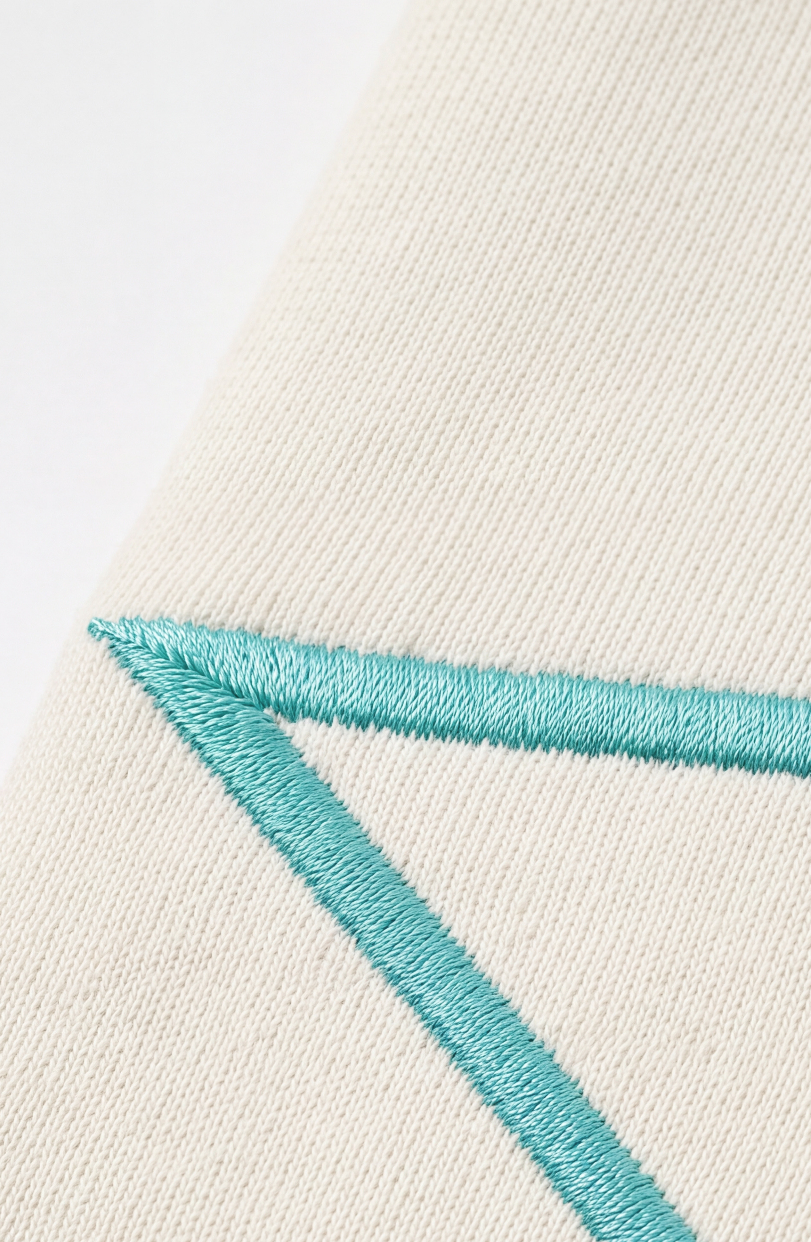 A close-up of cream cotton jersey fabric with blue-green geometric embroidery from the LOULOU Embroidered Star Sweatpants, showcasing intersecting lines and edges on a textured surface reminiscent of turquoise star motifs.